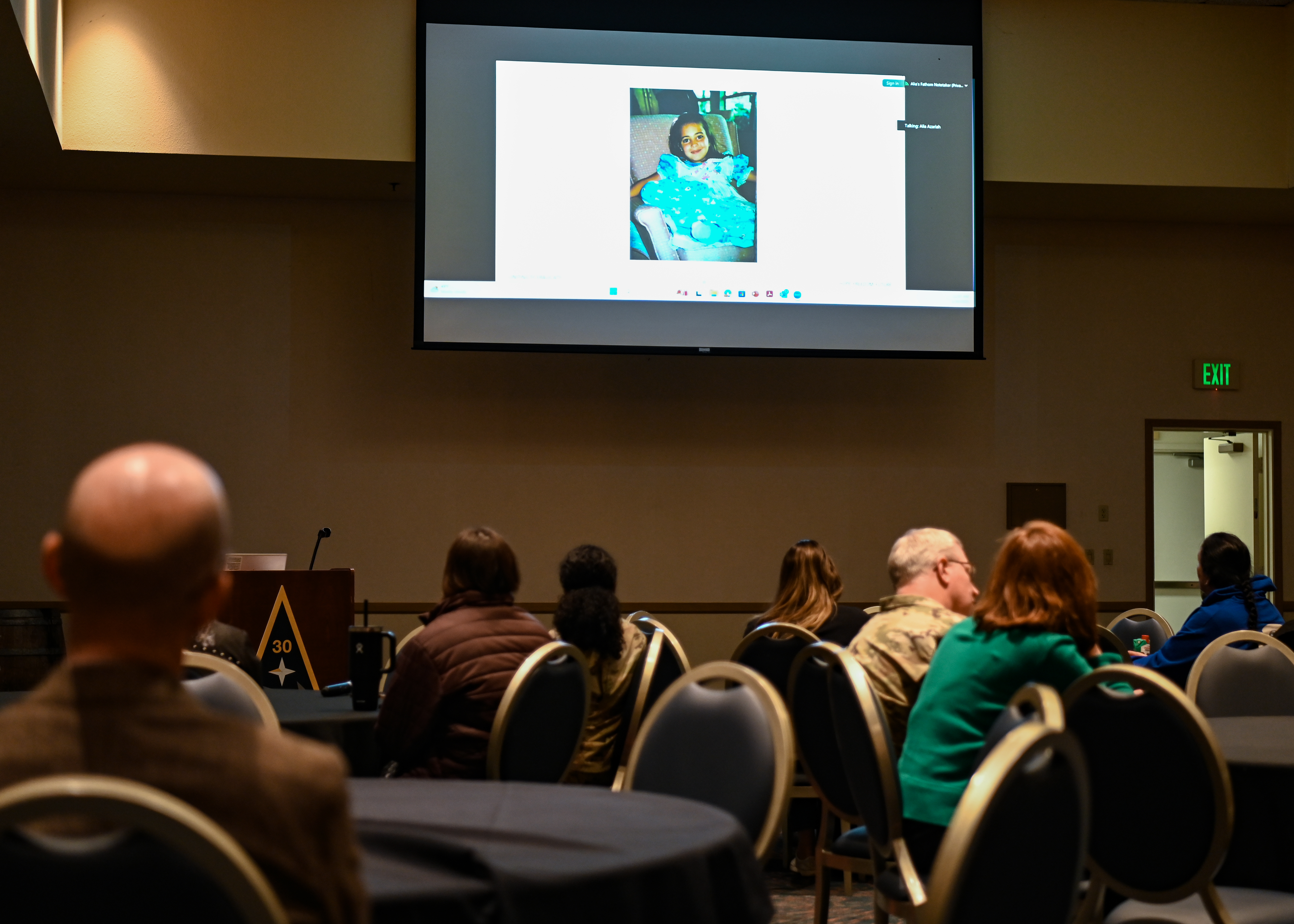 Audience members seated in rows watch a large projected screen showing a video call.