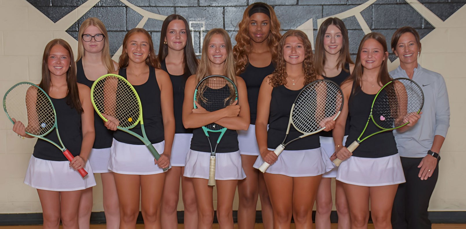 Group of young women in matching black tops and white skirts holding tennis rackets indoors.