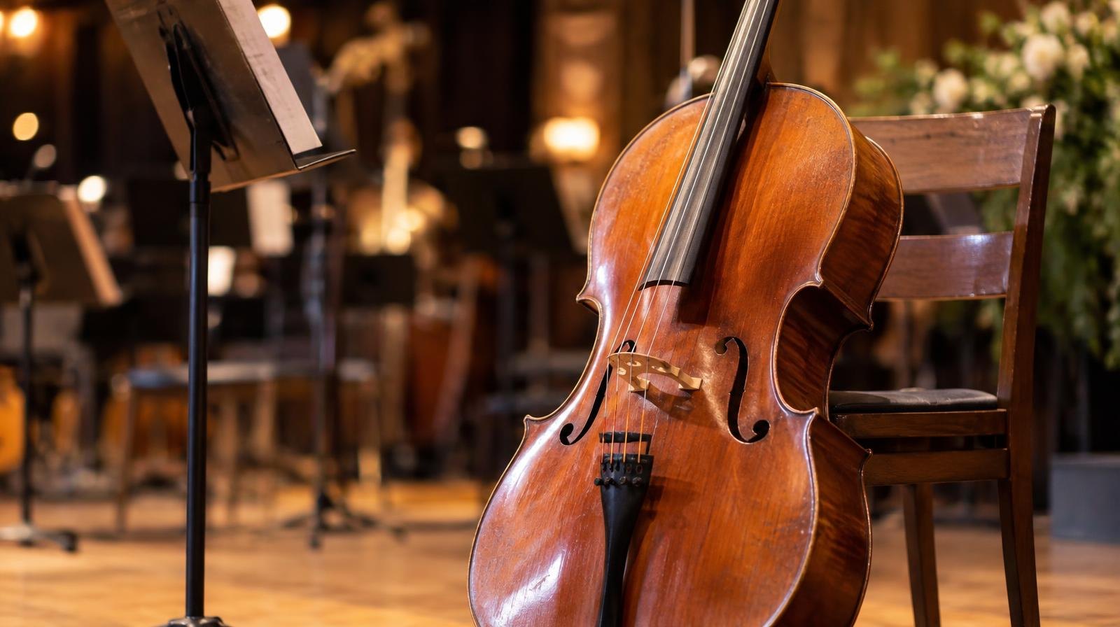A cello leaning against a wooden chair on a warmly lit concert stage.