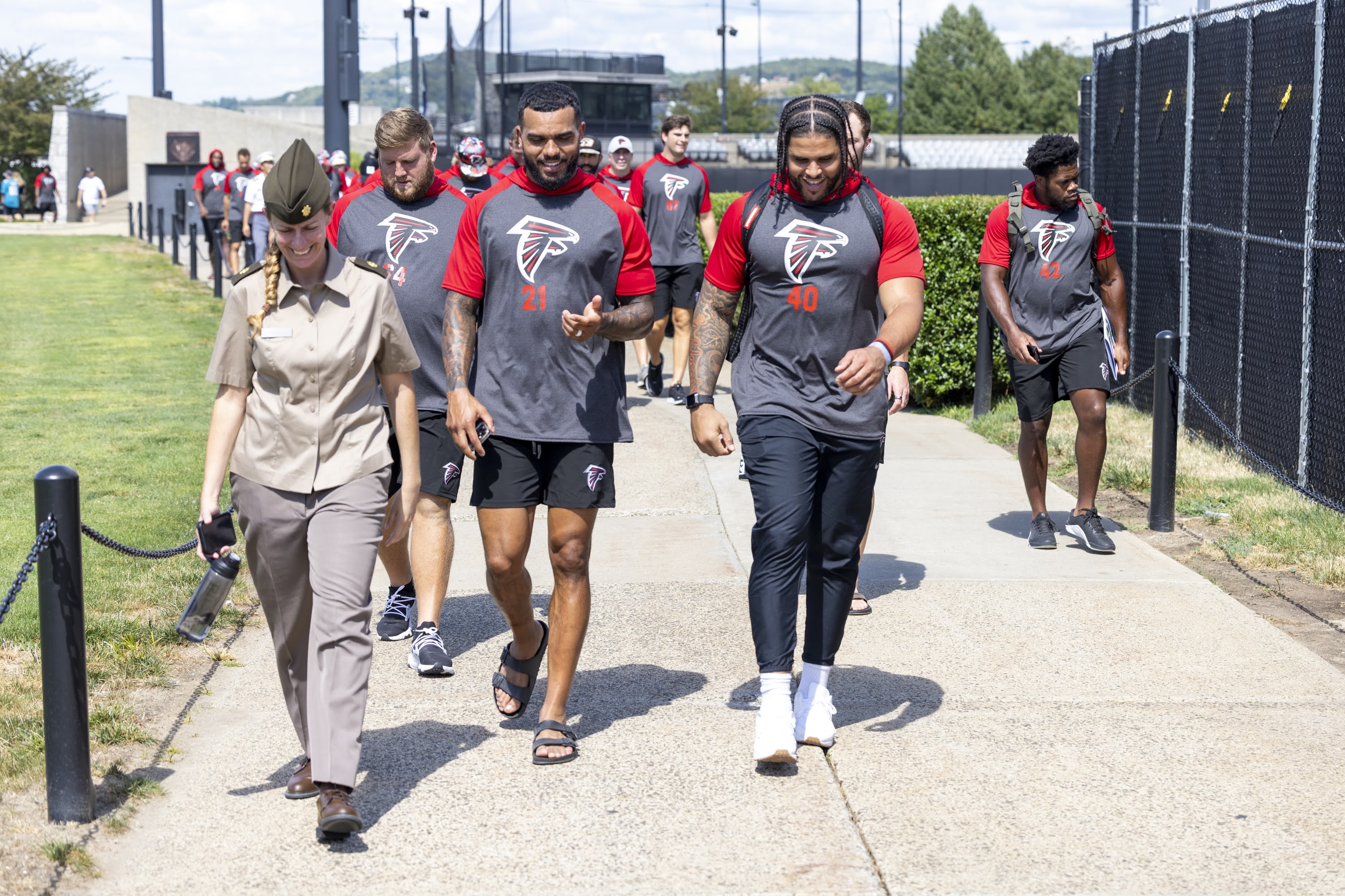 Group of football players in gray team shirts walking along a sidewalk with a uniformed military officer.