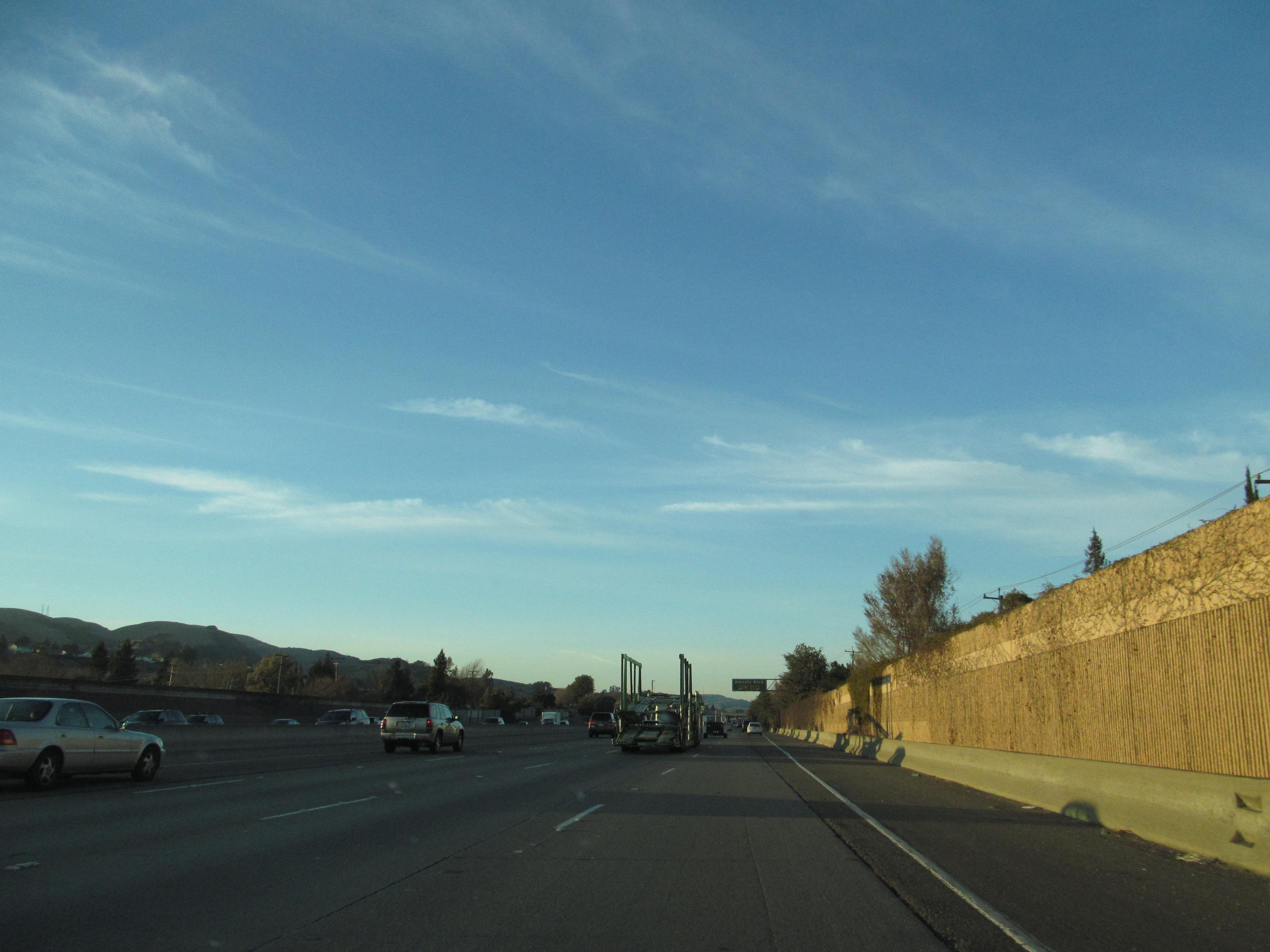 Multi-lane freeway with light traffic, a retaining wall on the right, and hills in the background.