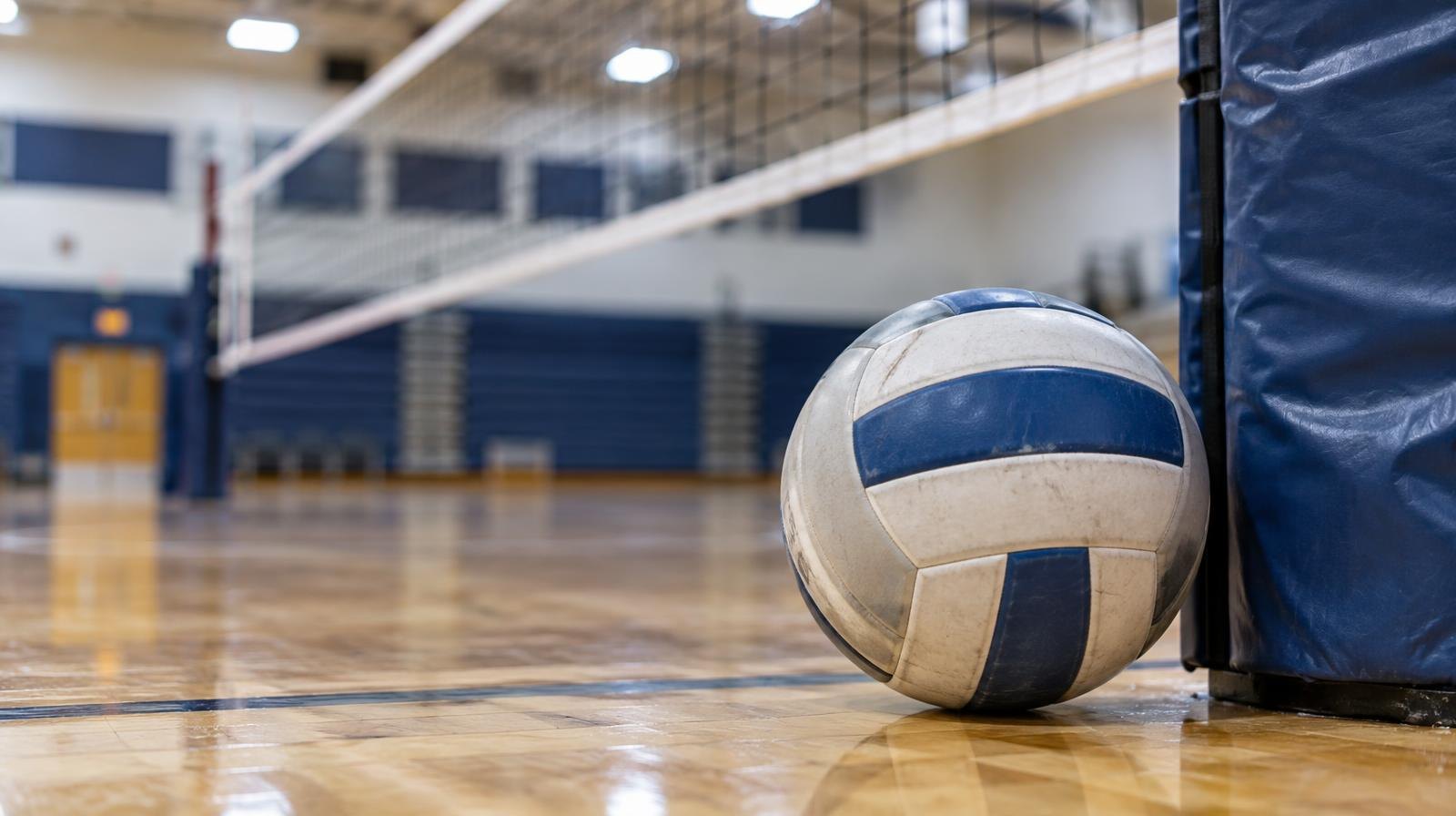 A volleyball resting on a hardwood gymnasium floor beside a net post.