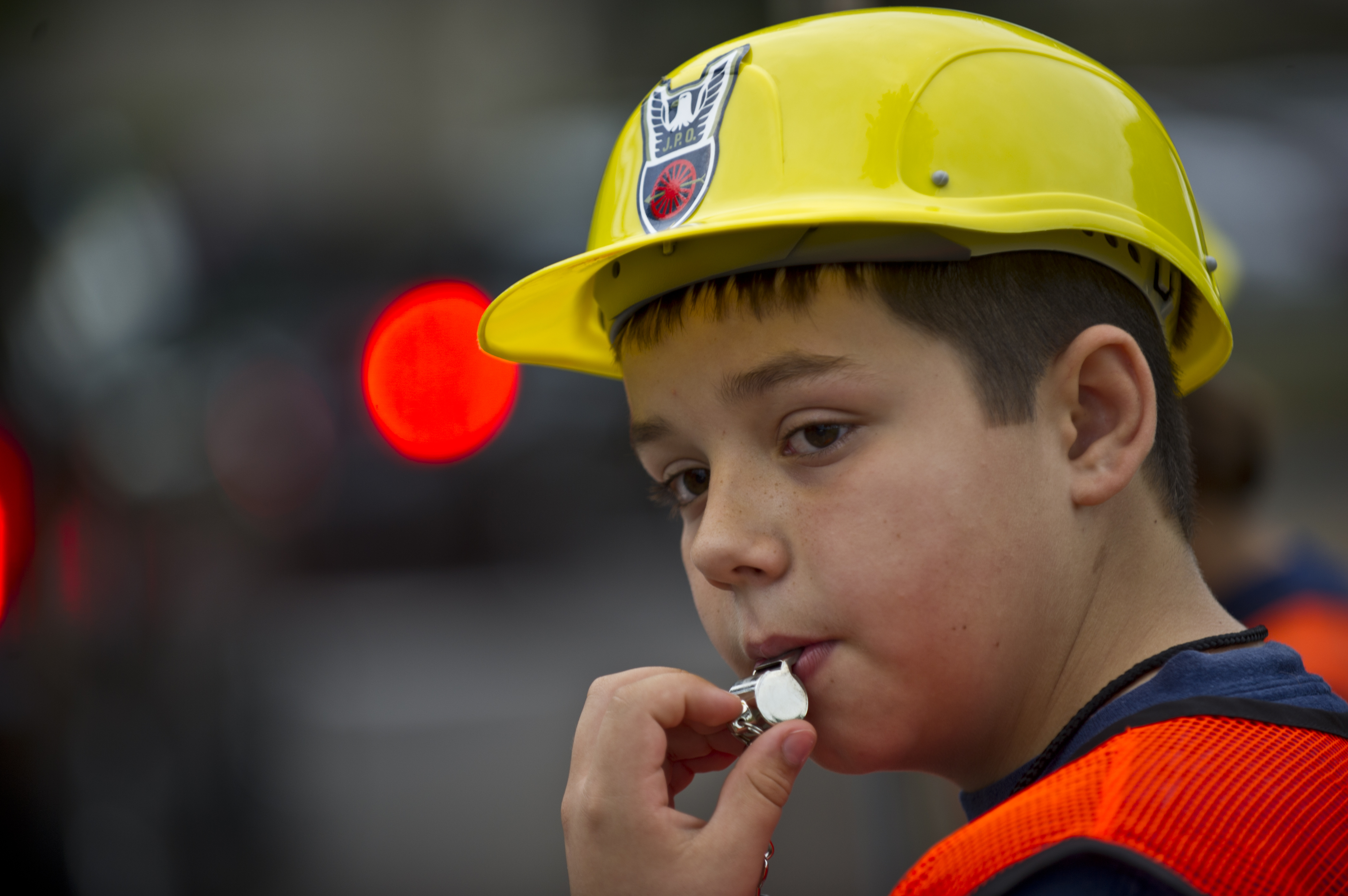 Child in orange safety vest and yellow hard hat blowing a whistle at a crosswalk.