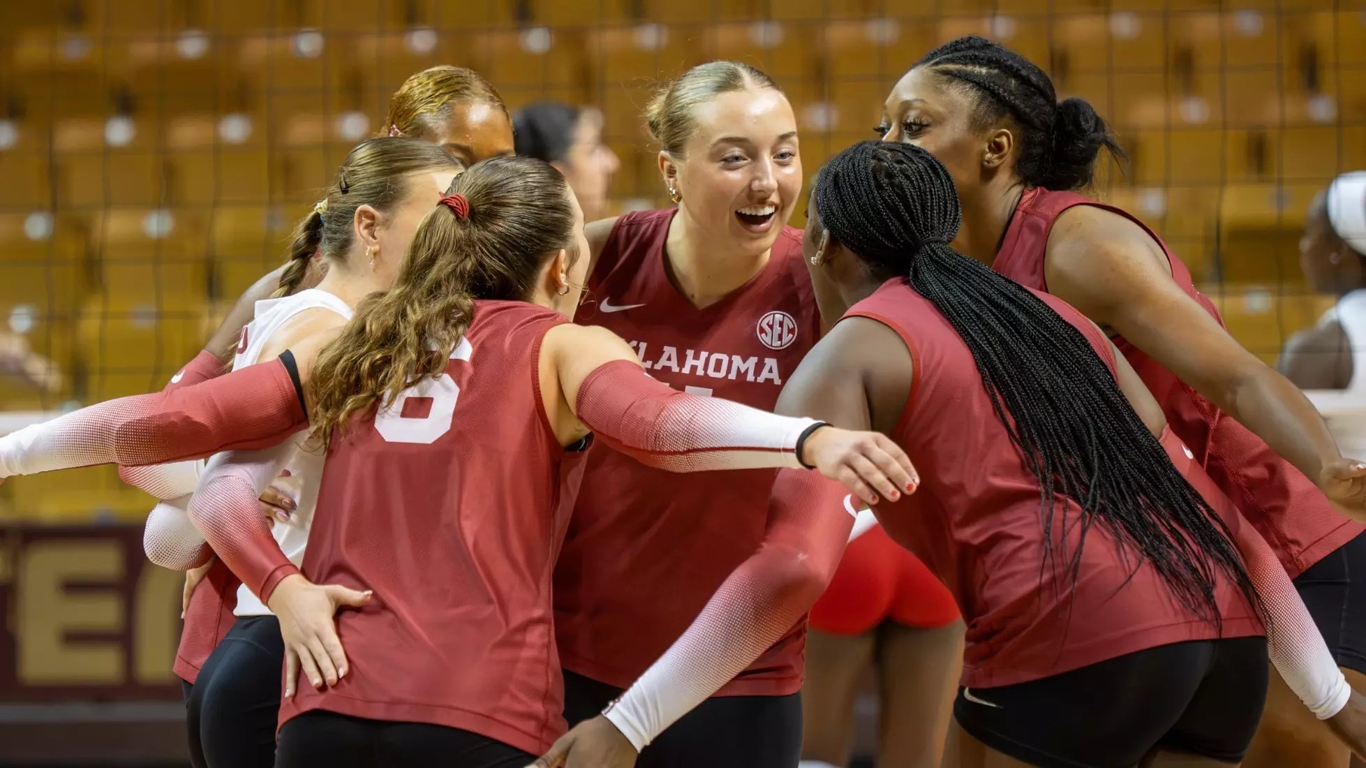 Female volleyball players in red Oklahoma uniforms embrace and celebrate on an indoor court.