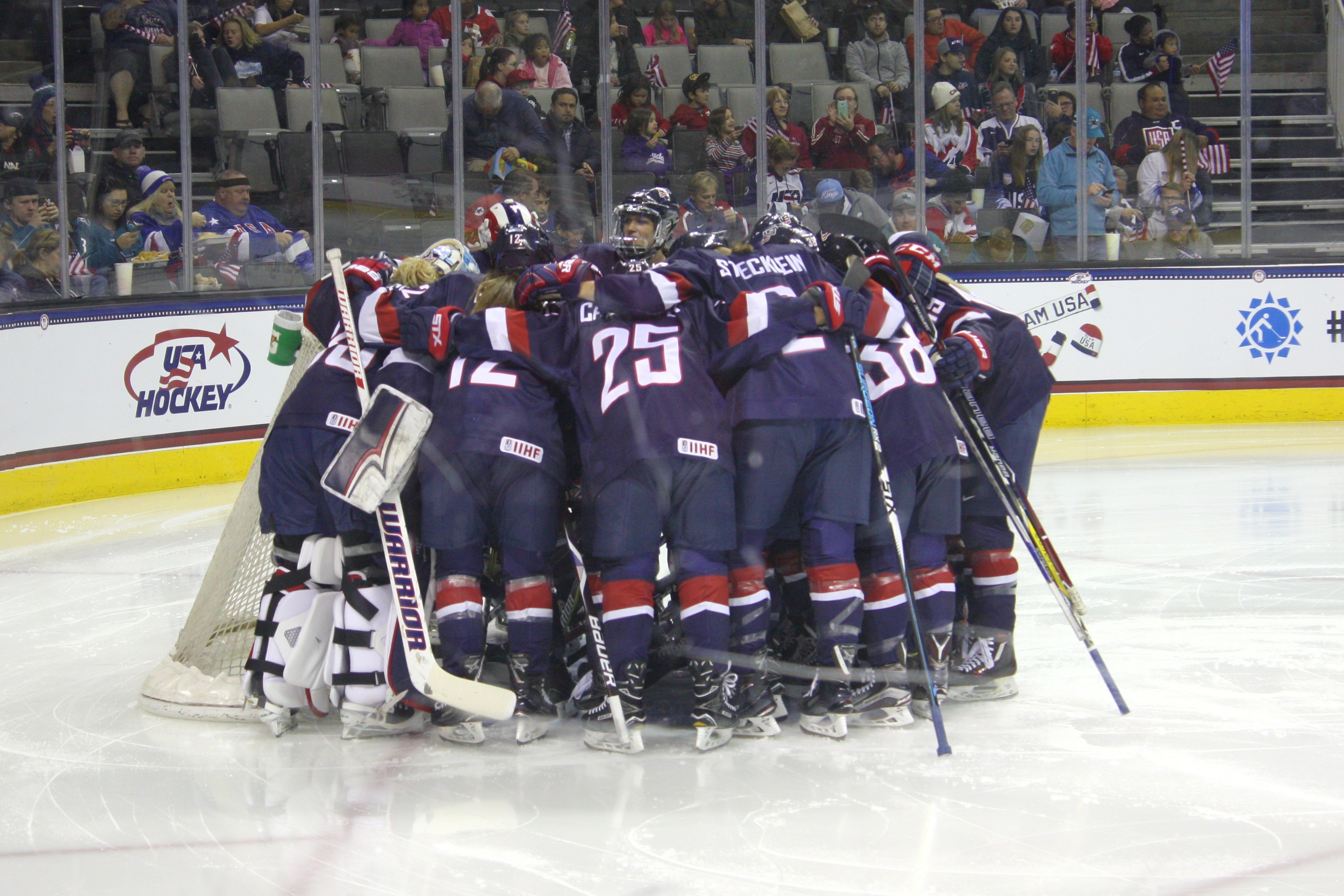 Women's hockey players in navy Team USA uniforms huddle near their goal on the ice.