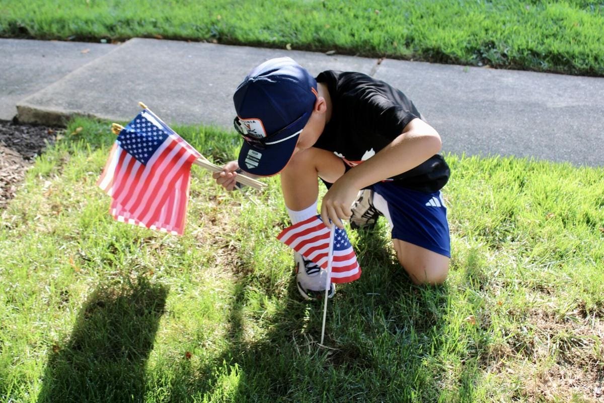 A boy in a navy cap kneels on grass planting small American flags near a sidewalk.