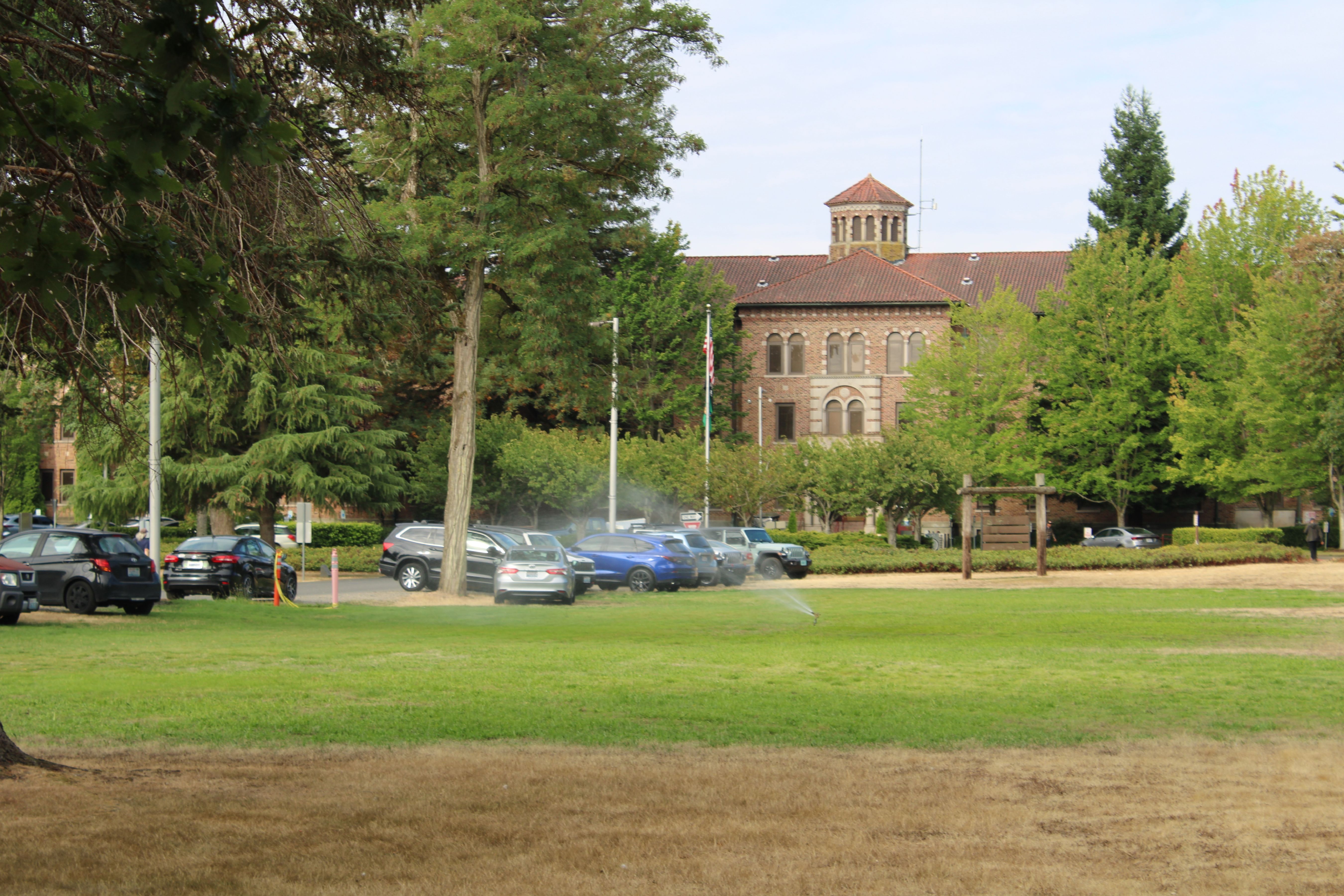 Large brick institutional building with bell tower behind a tree-lined parking lot.
