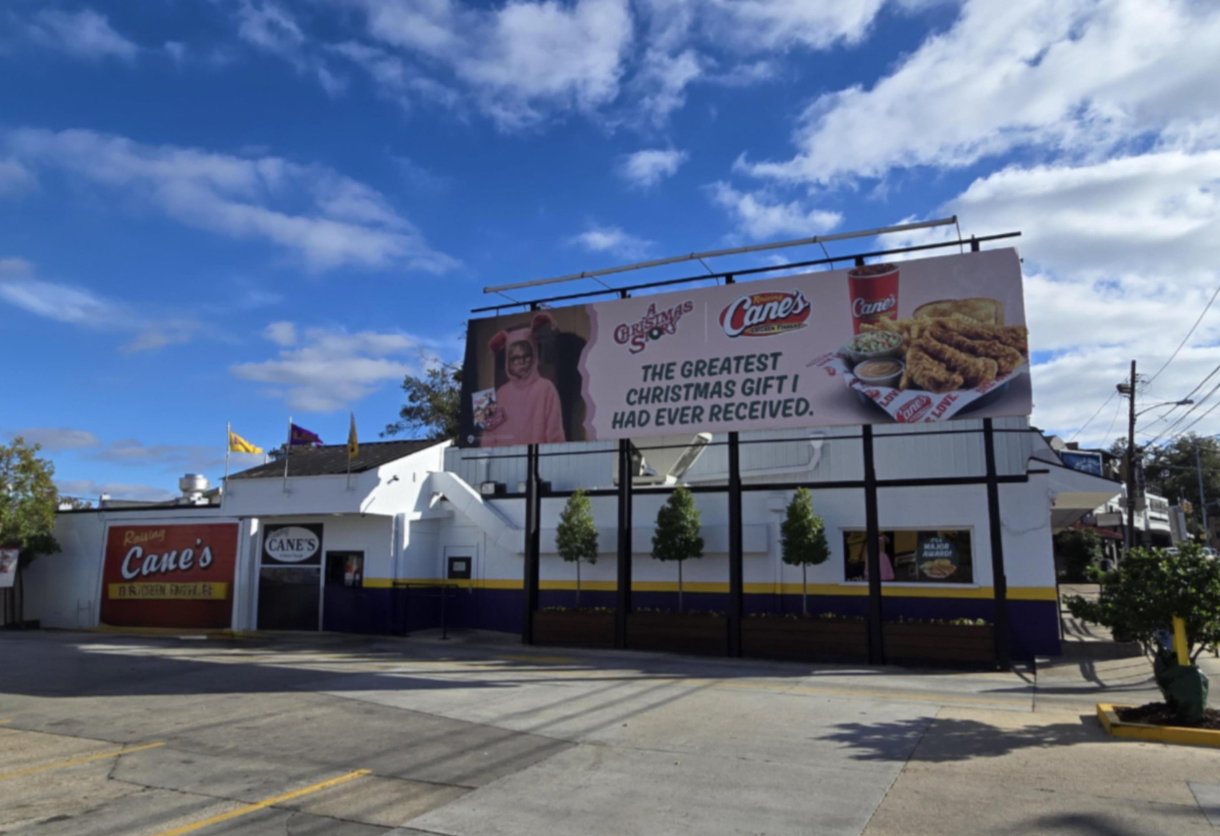 Fast food restaurant exterior with drive-thru lane and large rooftop billboard under a partly cloudy sky.