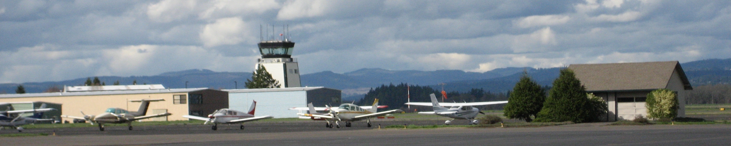 Airport tarmac with small planes, a control tower, hangars, and distant mountains.