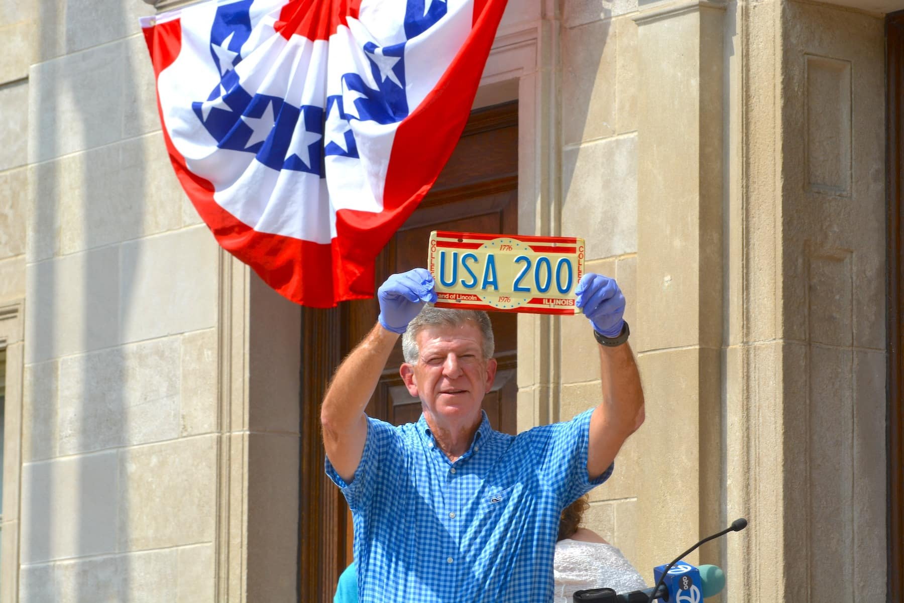Man in blue checked shirt holding up a USA 200 bicentennial license plate with patriotic bunting behind him.