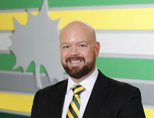 Bald man with brown beard in black suit and green-and-gold striped tie poses before a school-colored striped wall.