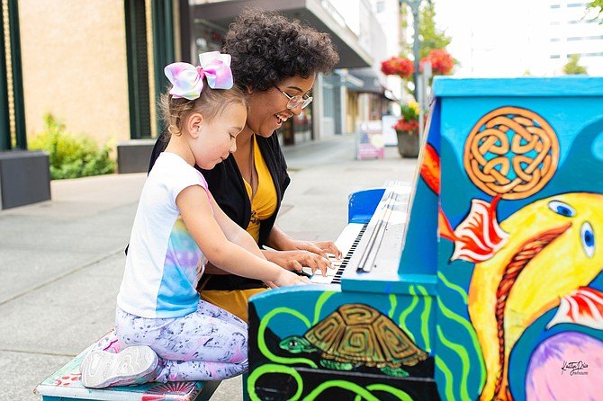 Two people play a brightly painted upright piano on an outdoor urban sidewalk.