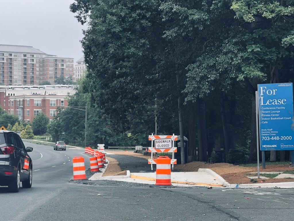 Newly paved sidewalk blocked by orange traffic barrels and a sidewalk closed sign along a tree-lined road.