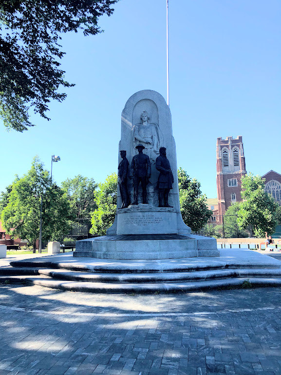 Stone war memorial with bronze figures stands in a park under a blue sky.