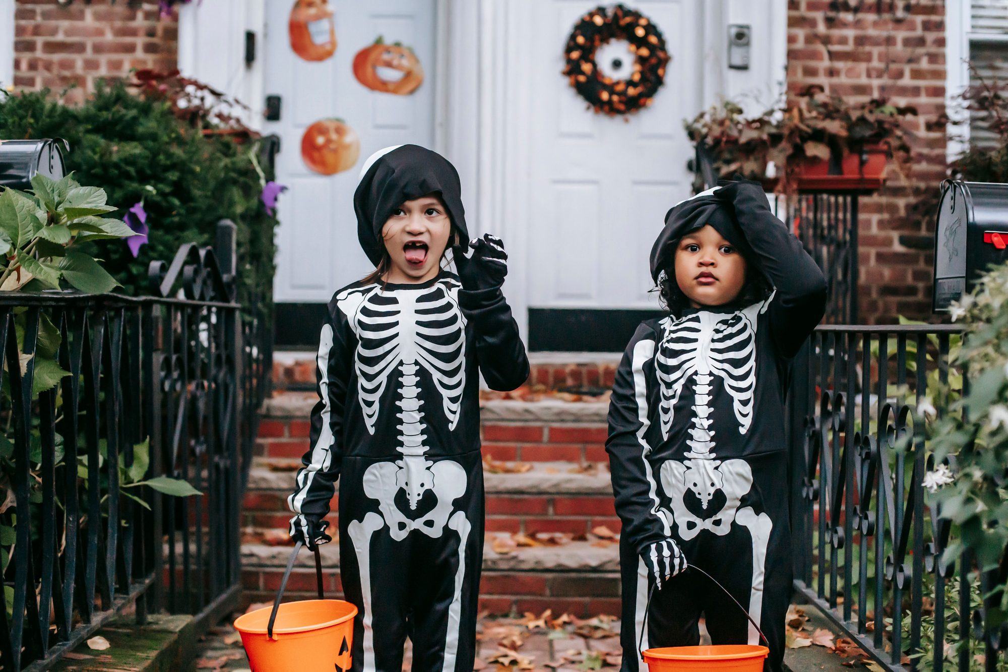Multiethnic girls in skeleton costumes with orange buckets standing near house door and grimacing in Halloween