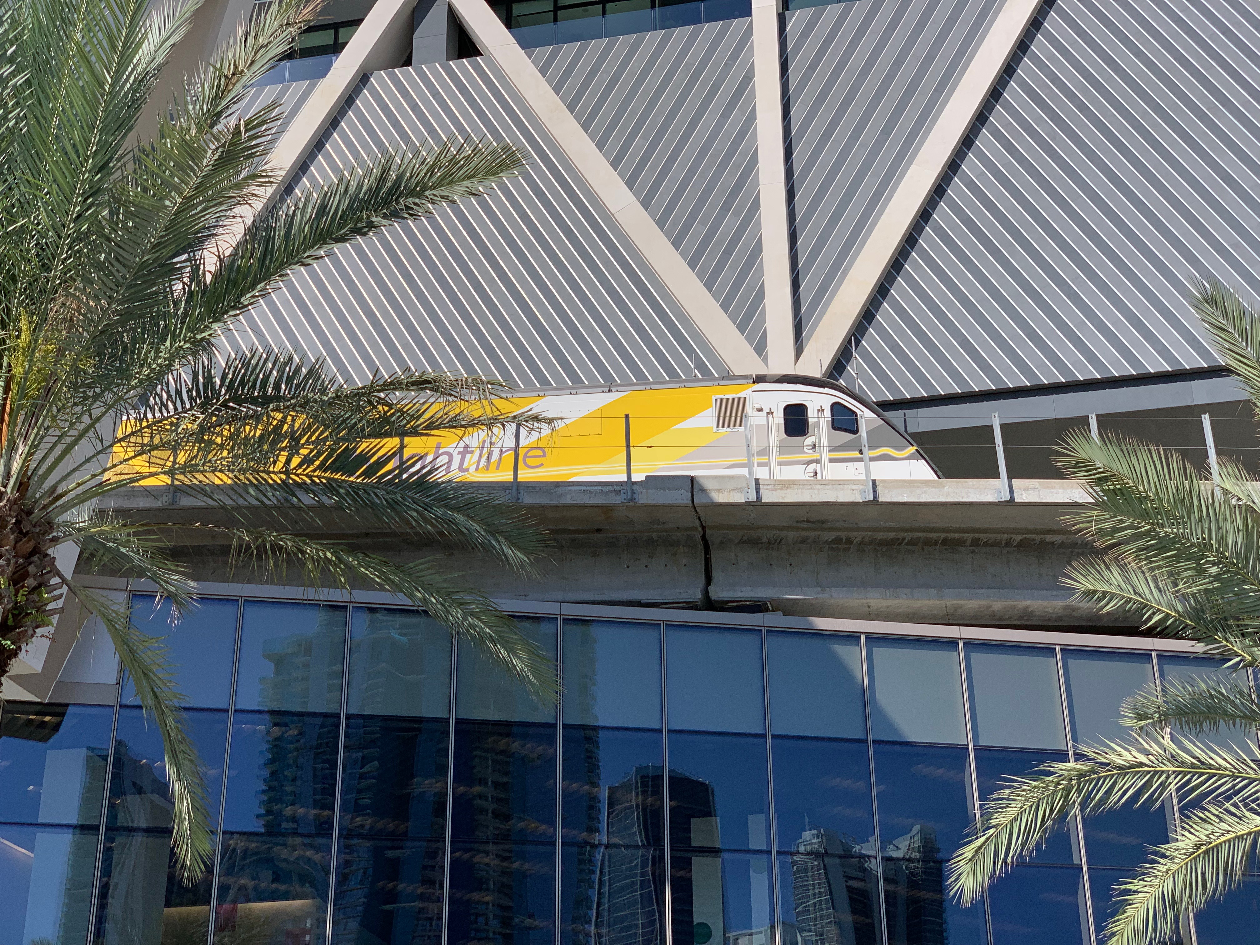 A yellow Brightline train on an elevated track beside a modern glass-and-steel station building.