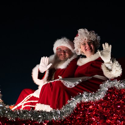Two people dressed as Santa and Mrs. Claus wave from a tinsel-decorated float at night.