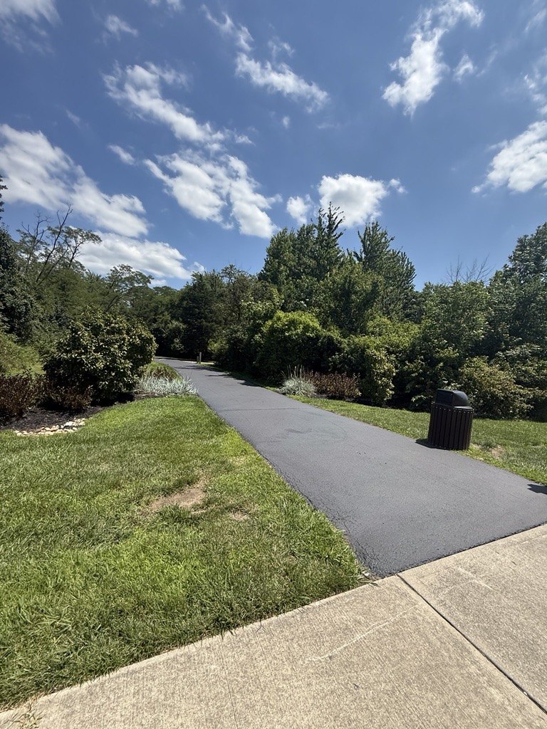 Newly paved dark asphalt trail winding through green landscaping and trees.