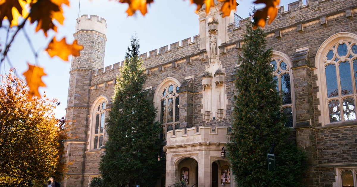 Gothic stone academic building with crenellated towers and autumn foliage.