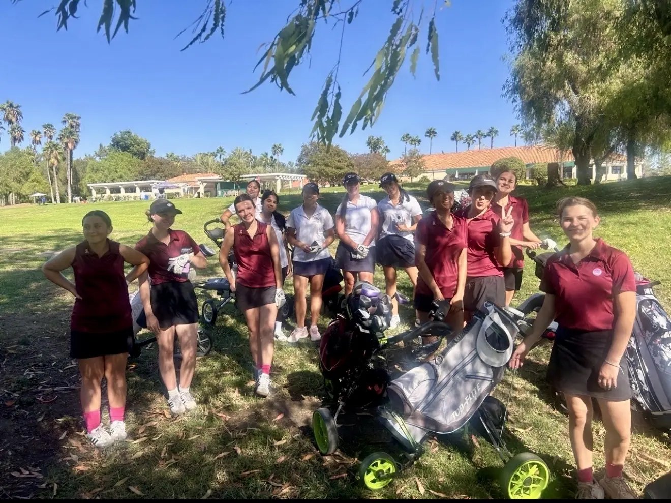 A group of young female golfers in matching polo shirts pose together on a golf course with bags and pull carts.