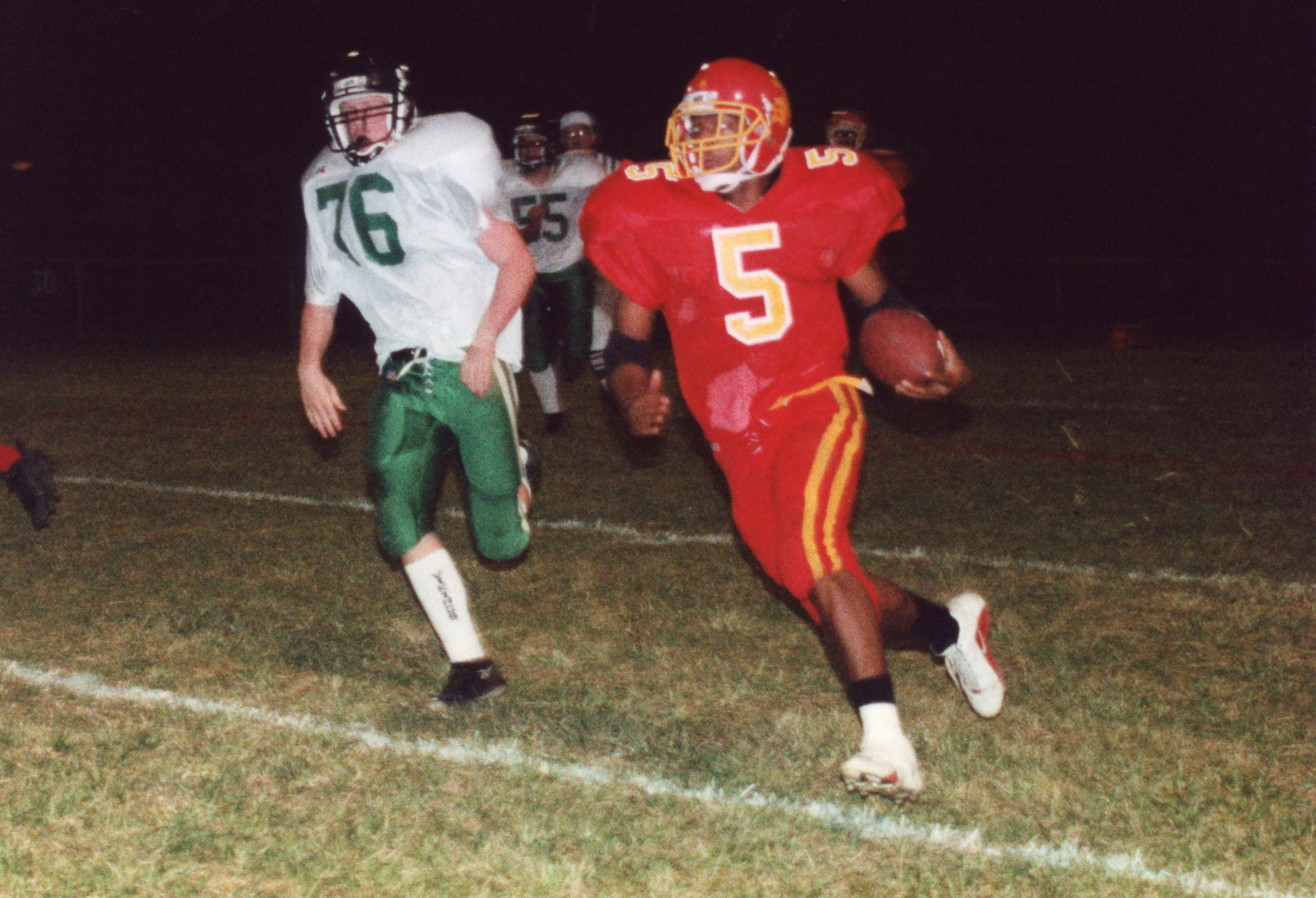 A high school football player in a red jersey runs with the ball at night.