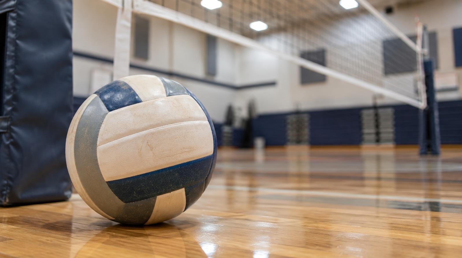 A volleyball resting on a hardwood gymnasium floor near the net post.