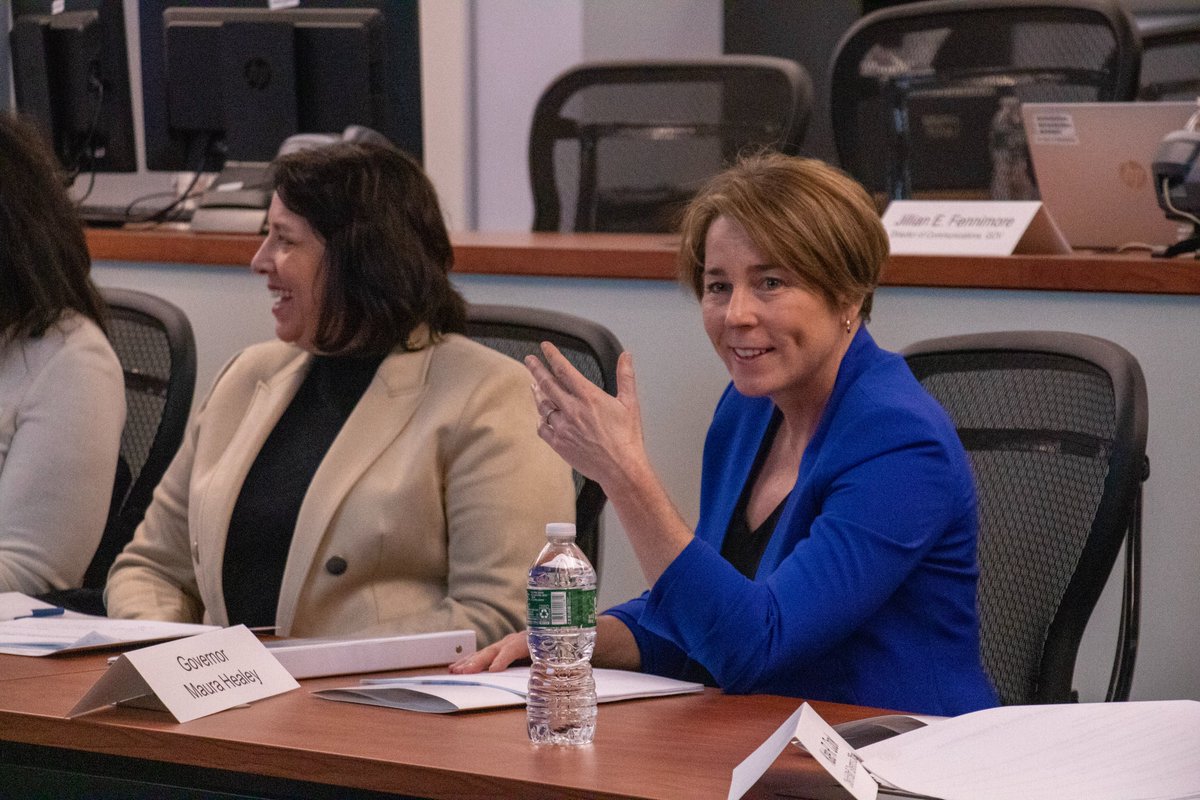 A woman in a blue blazer gestures at a conference table bearing a nameplate reading 'Governor Maura Healey.'