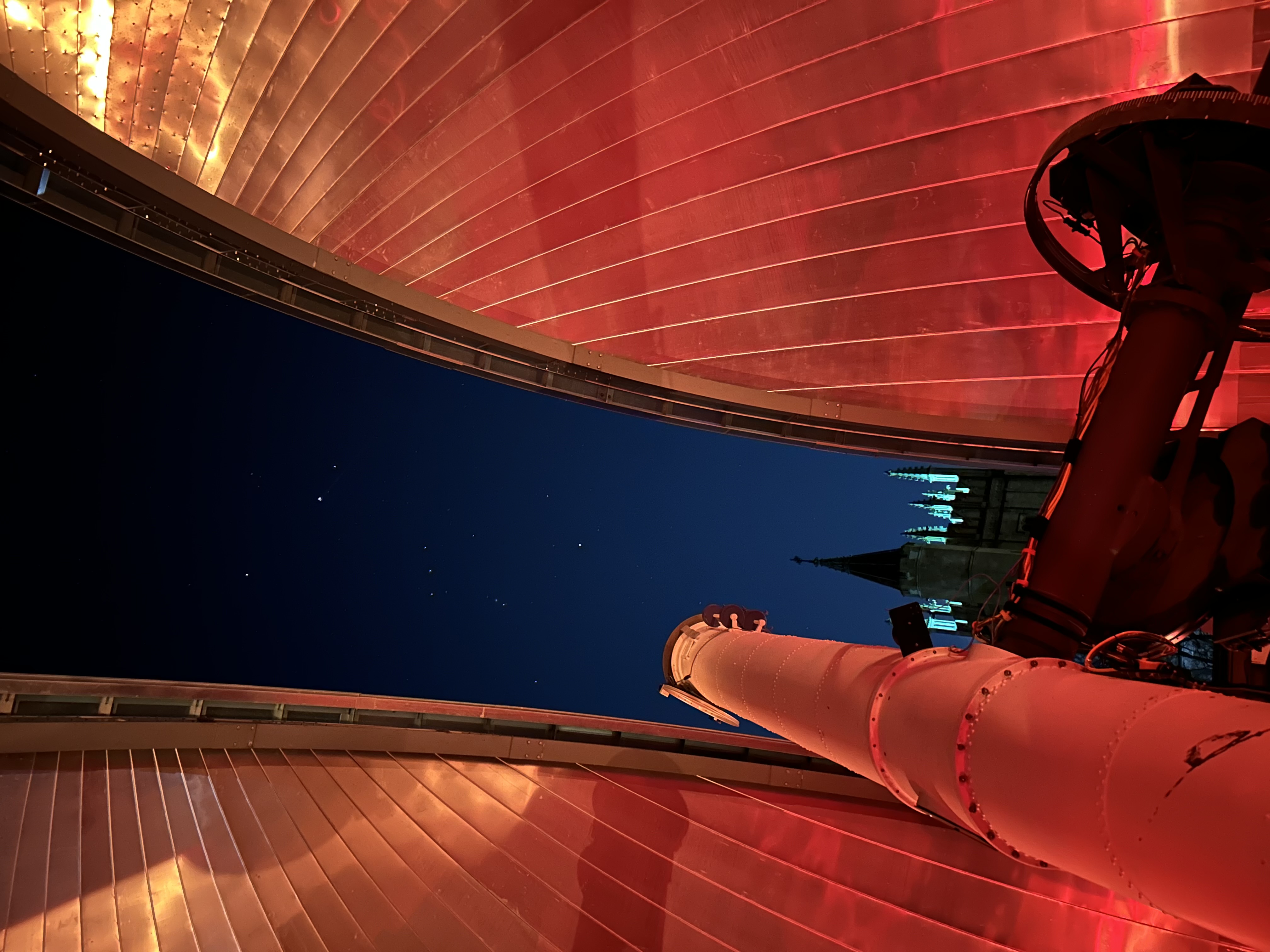 Large refracting telescope inside an observatory dome open to a starry night sky, lit by red lights.