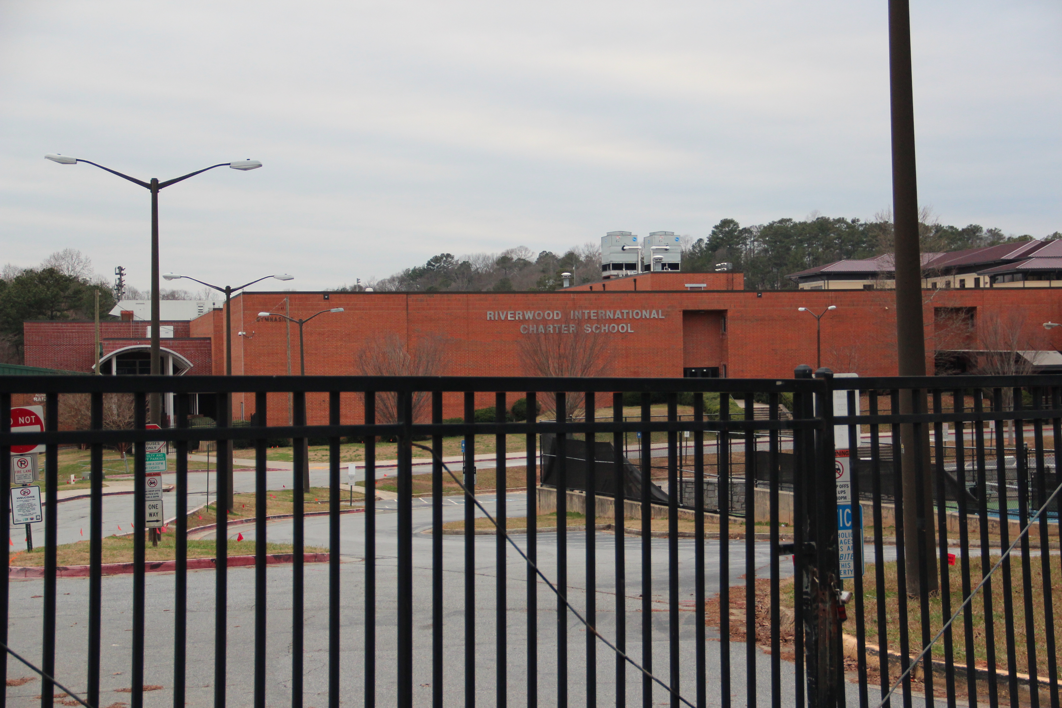 Brick school building seen through a black iron security fence in a parking lot.
