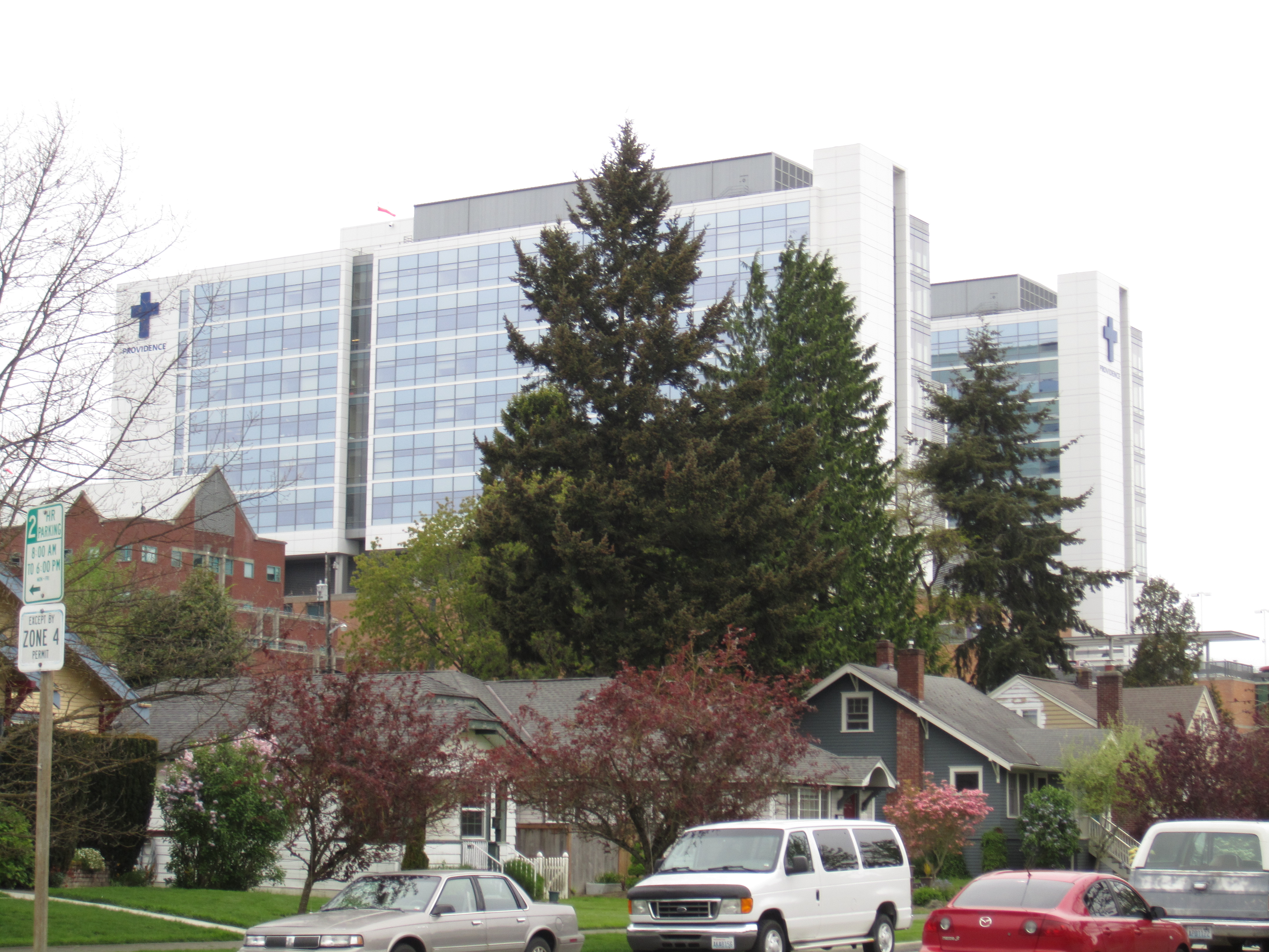 Tall glass-and-steel hospital tower with blue cross symbols, surrounded by trees and residential homes.