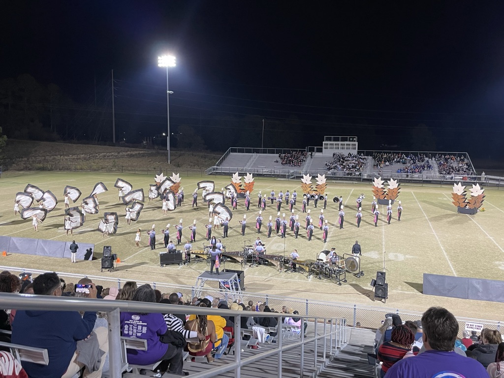 High school marching band performing on a football field at night with colorguard flags.