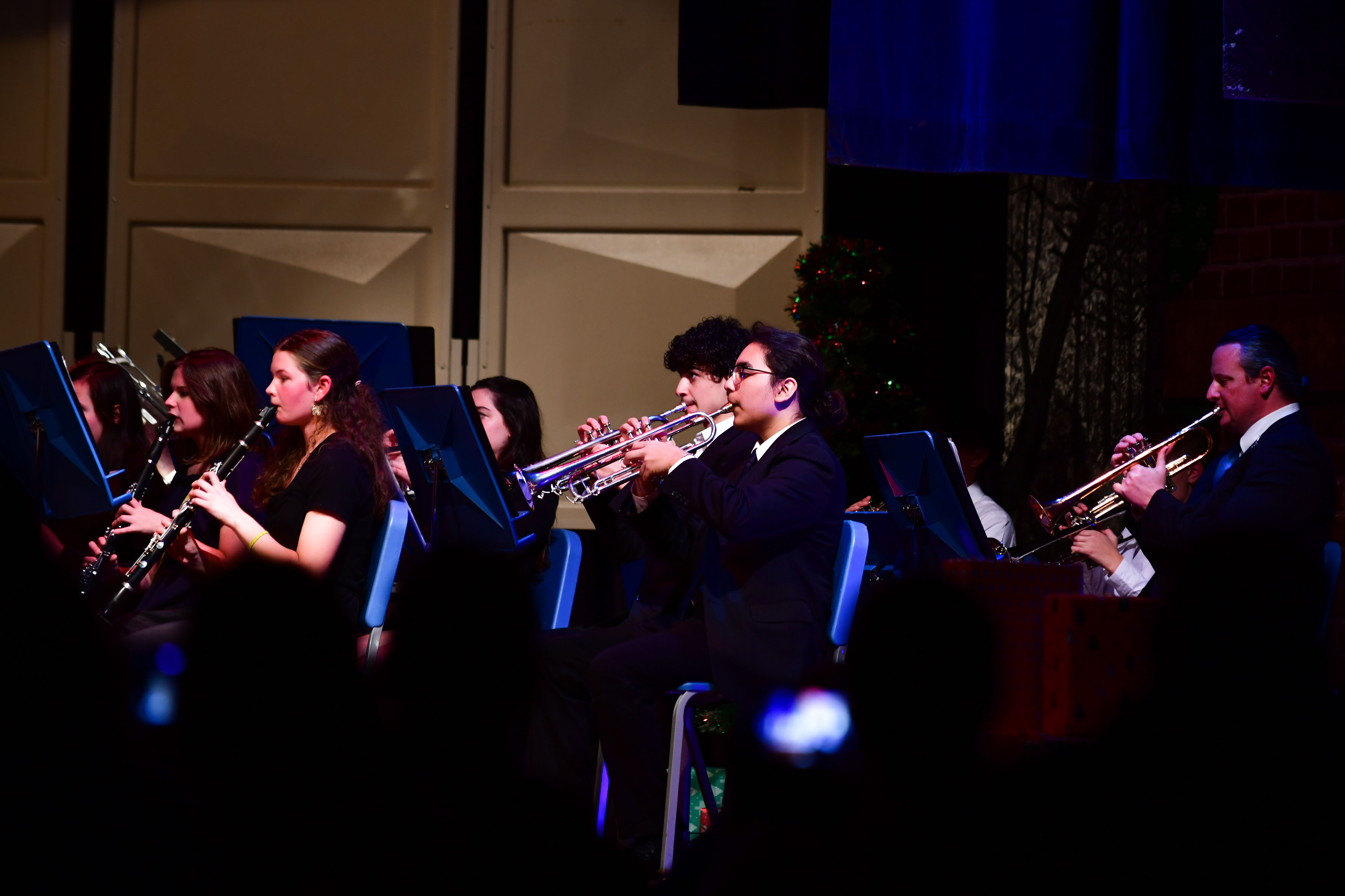 Students in black formal attire play wind instruments on a concert stage under blue lighting.