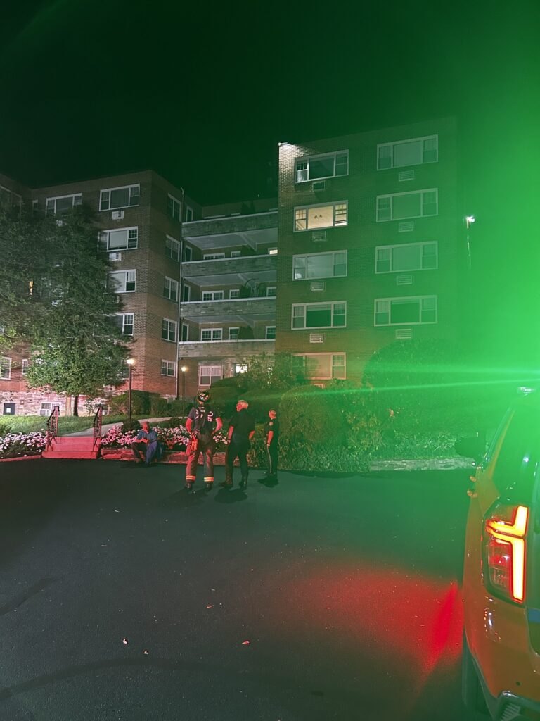 Firefighters in dark uniforms stand outside a tall brick apartment building at night.