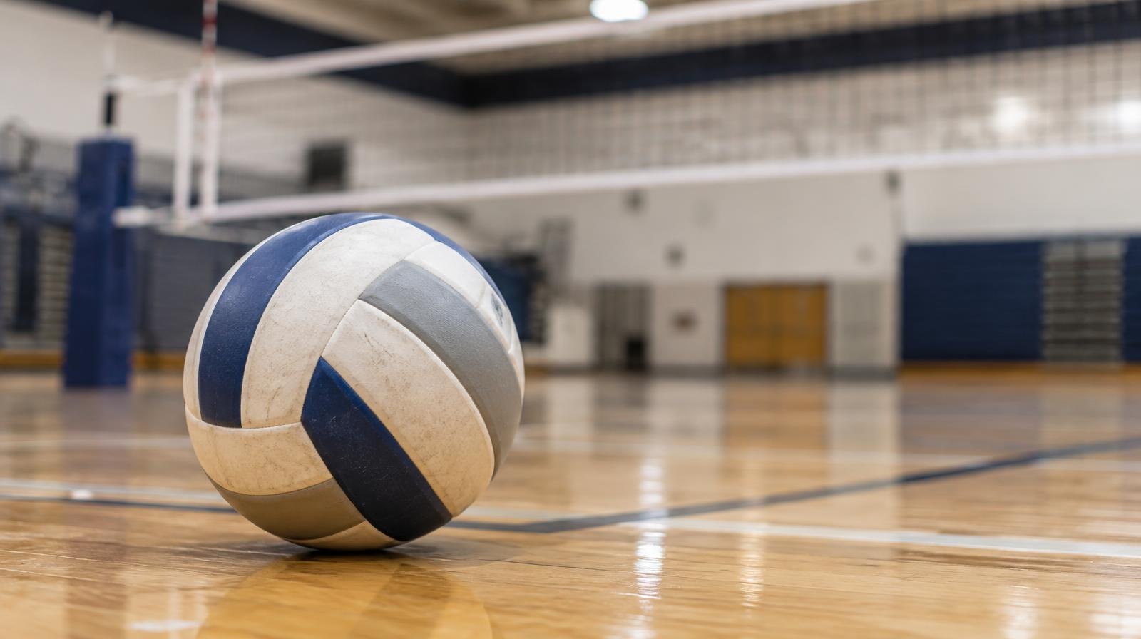 A volleyball resting on a hardwood floor near a volleyball net.