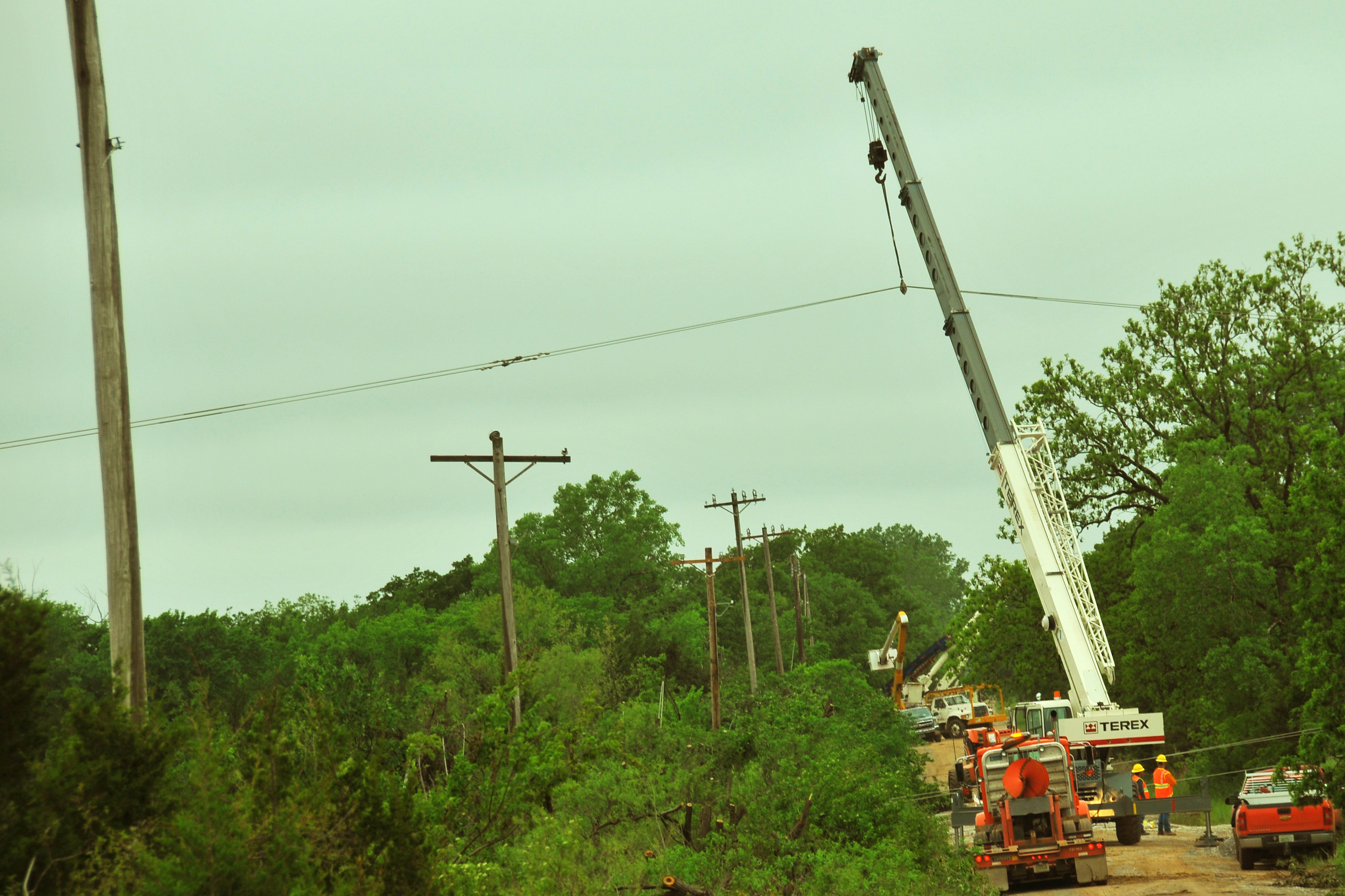 Utility workers and a crane replacing downed power lines on poles along a tree-lined road.