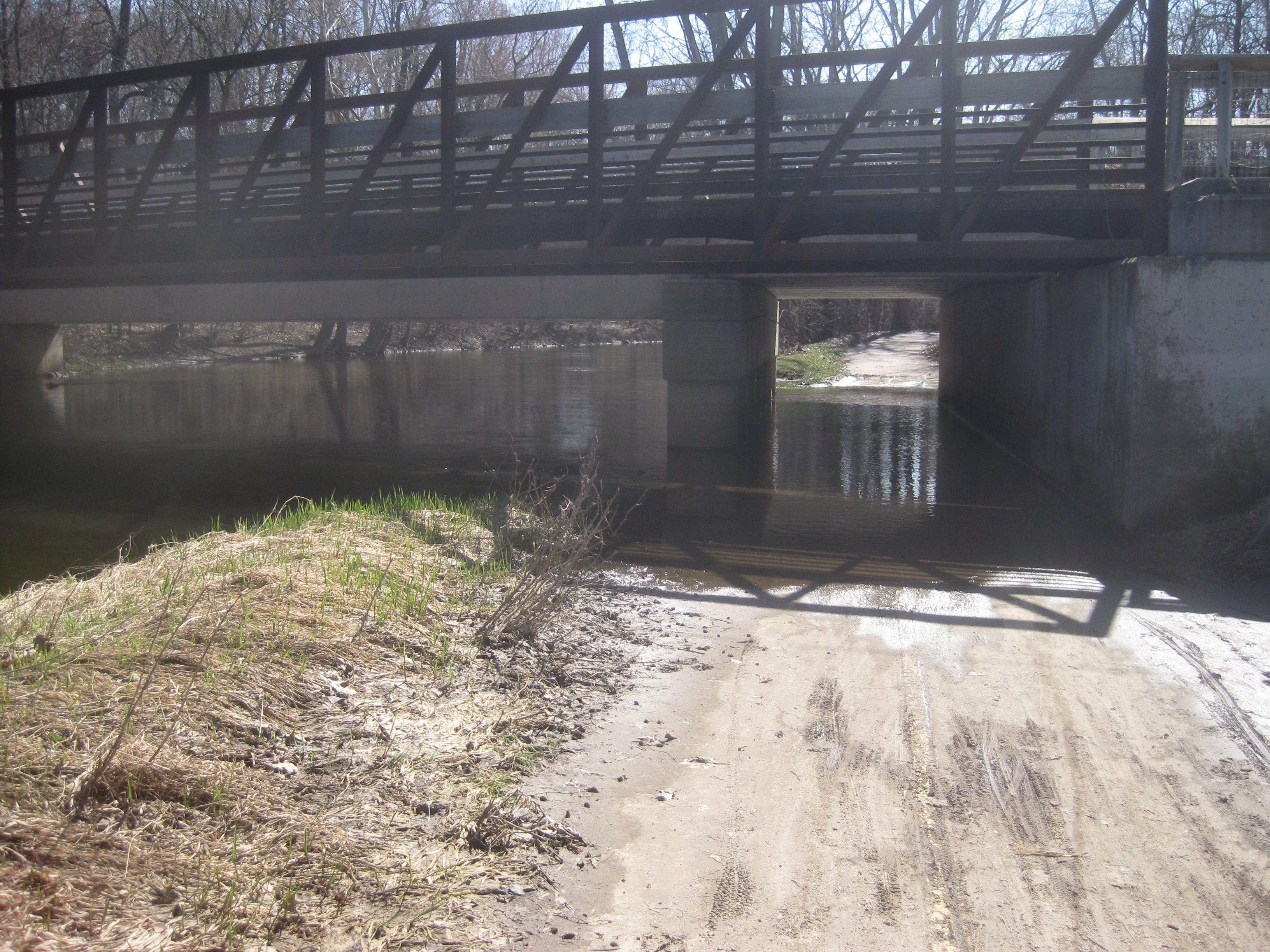 A flooded riverside trail beneath a steel truss bridge, with bare trees and sandy path.