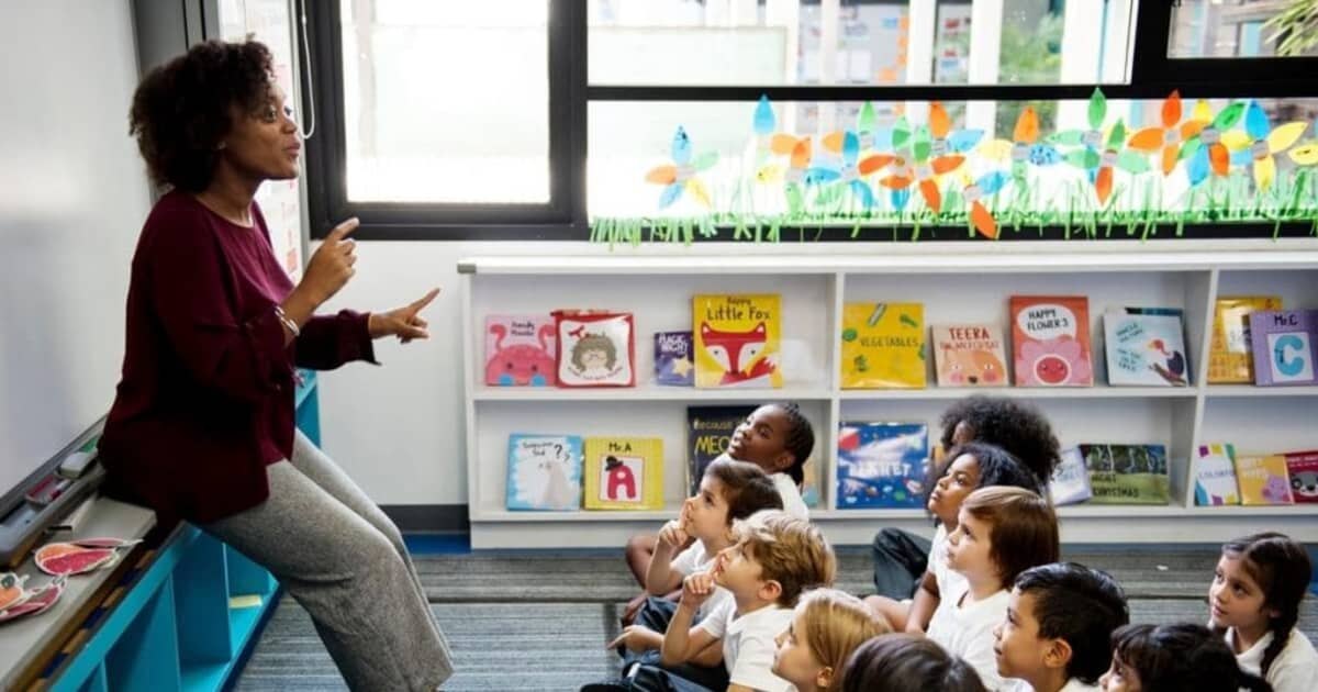 A female teacher speaks to young children seated on a classroom floor.