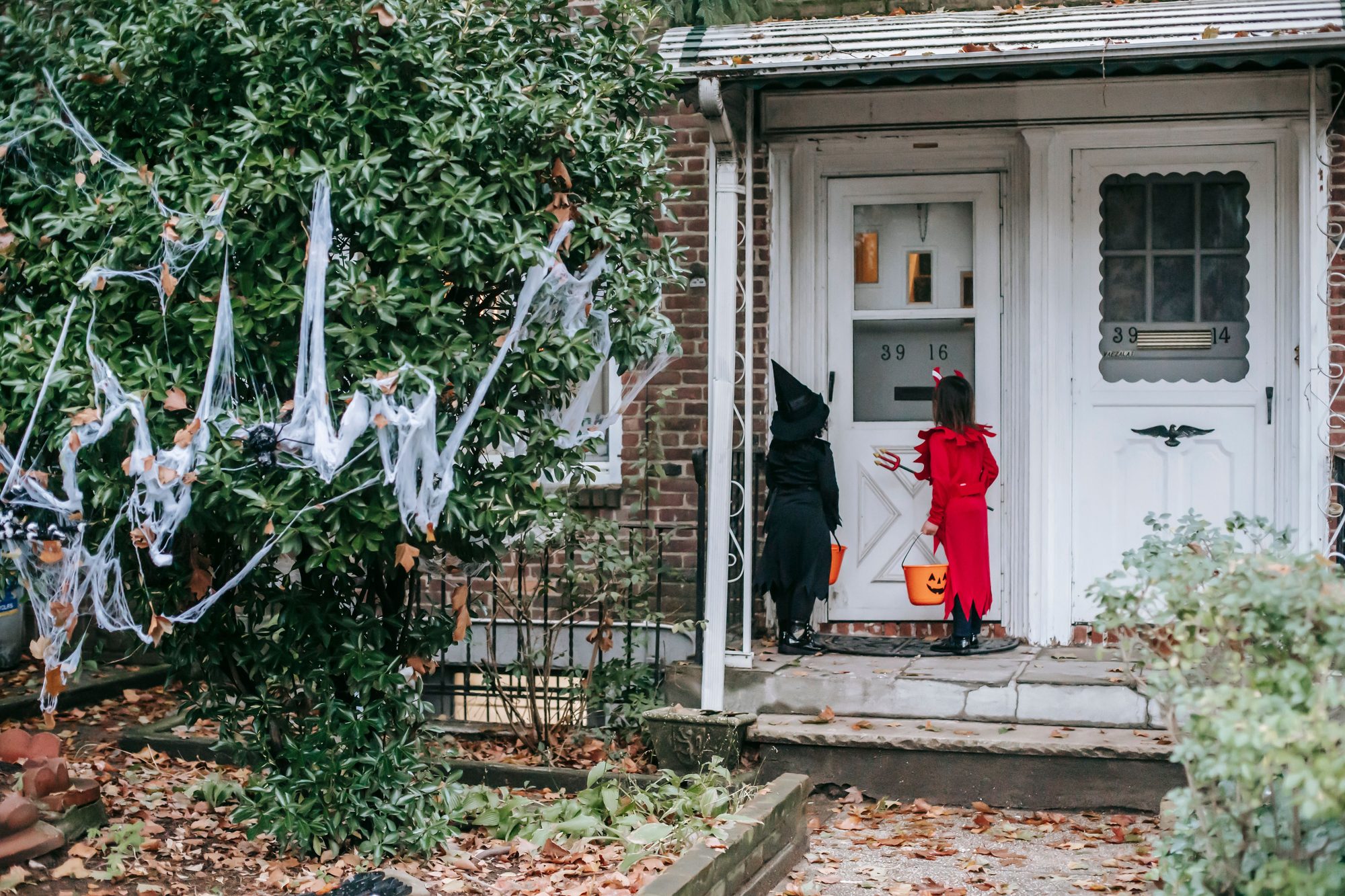 Full body back view of anonymous children in witch and devil costumes with buckets standing near entrance of house while playing trick or treat