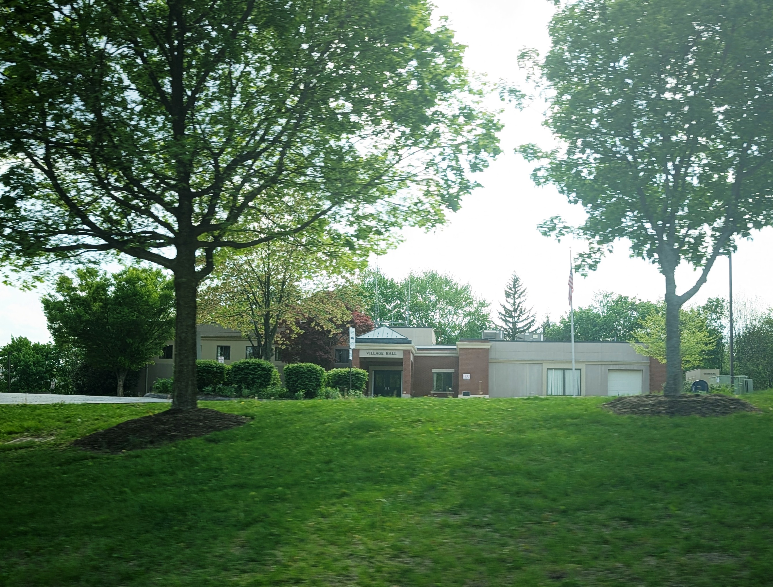 A low-rise municipal building with brick and light-colored siding surrounded by a manicured lawn and large trees.