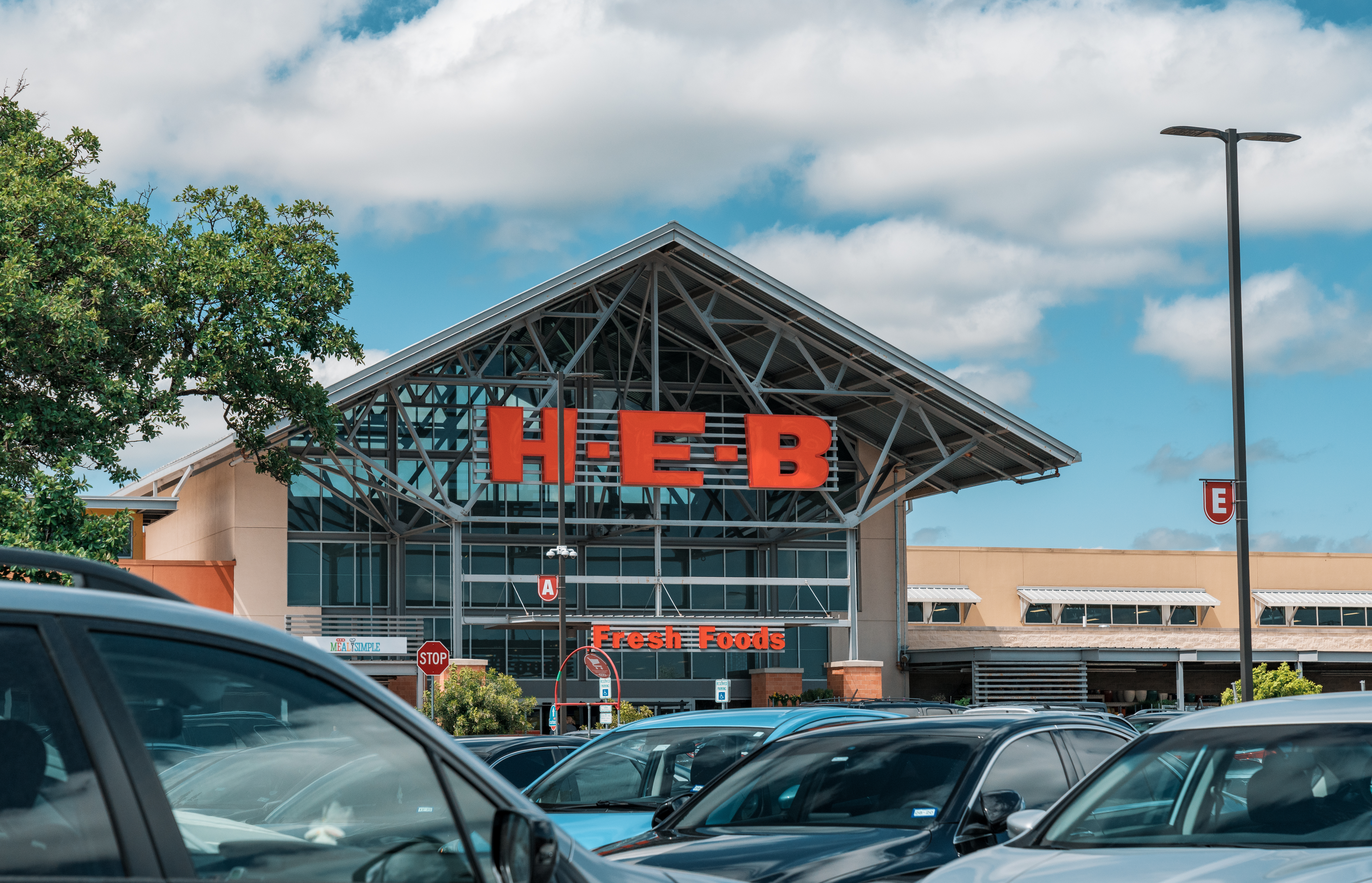 Exterior of an H-E-B grocery store with large orange signage and a full parking lot.