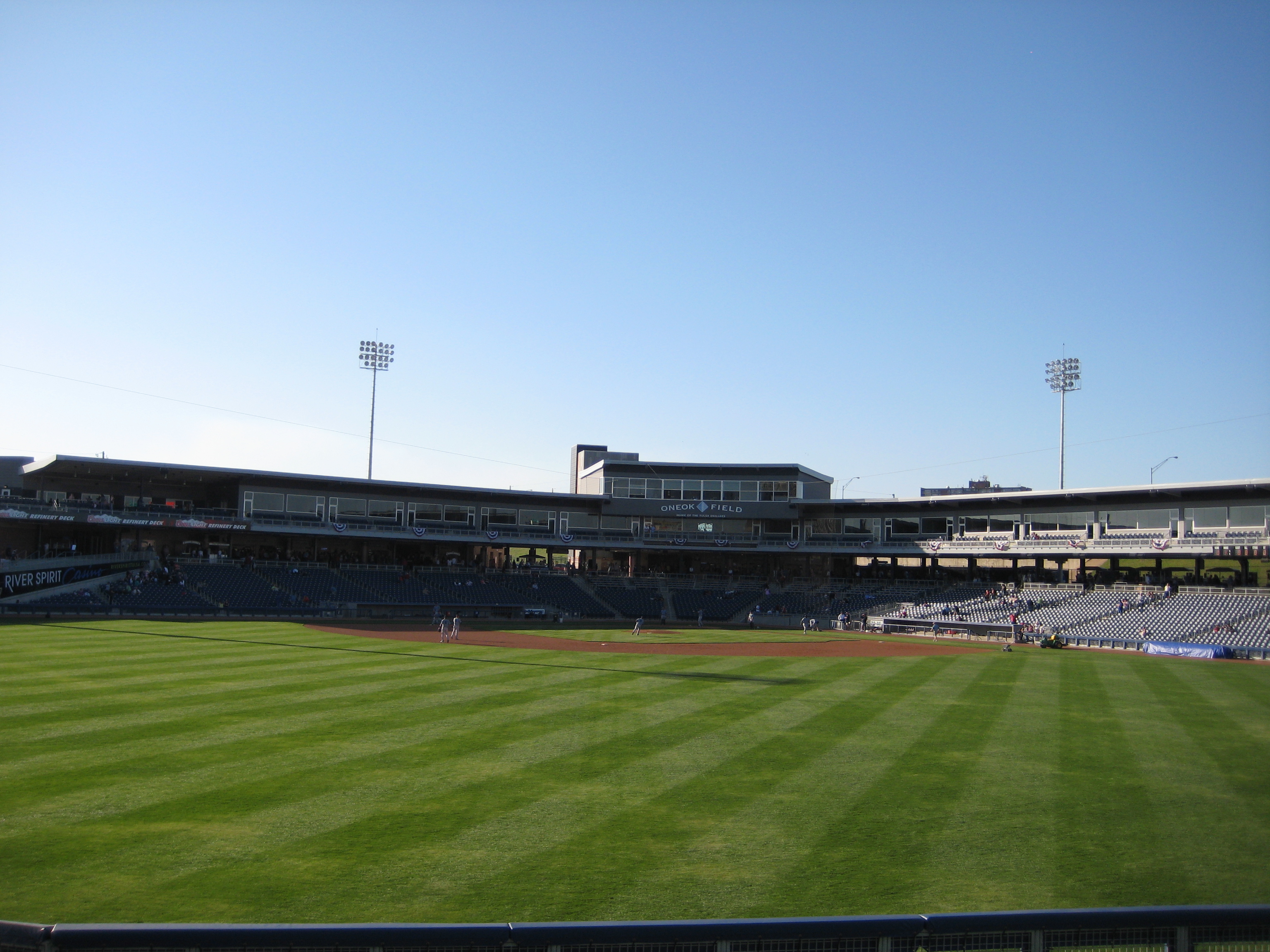 Minor league baseball stadium viewed from left-center field, showing grandstand and light towers under a blue sky.