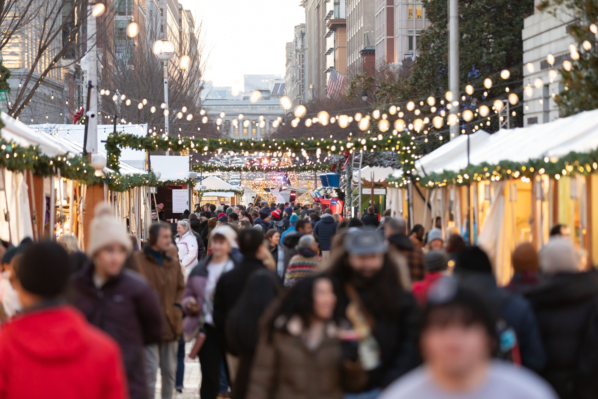 Crowds of shoppers walk through an outdoor holiday market lined with white vendor stalls and string lights.