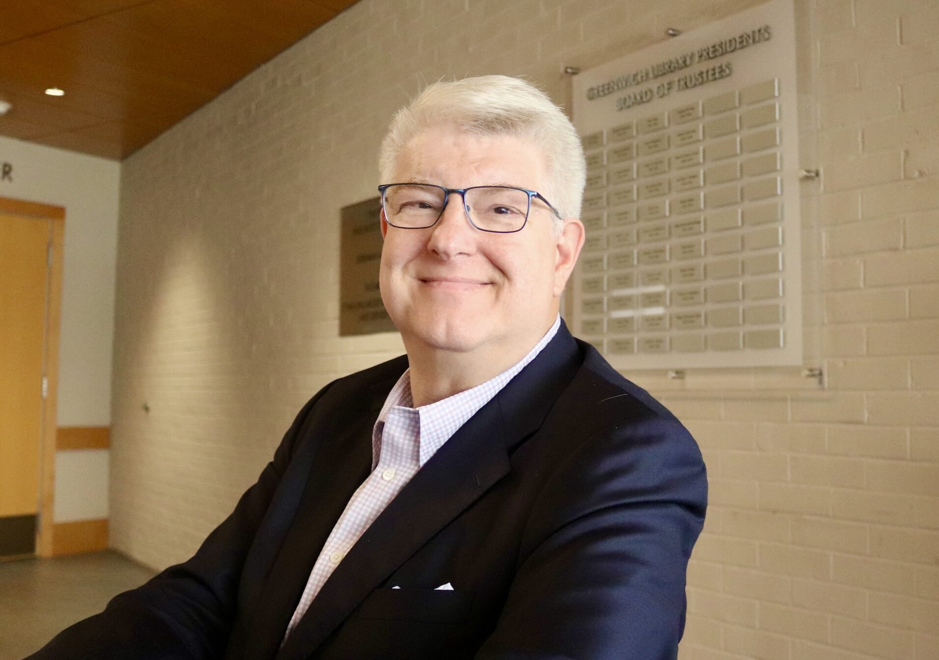 A smiling man with white hair and glasses wearing a dark blazer stands in a public building hallway.
