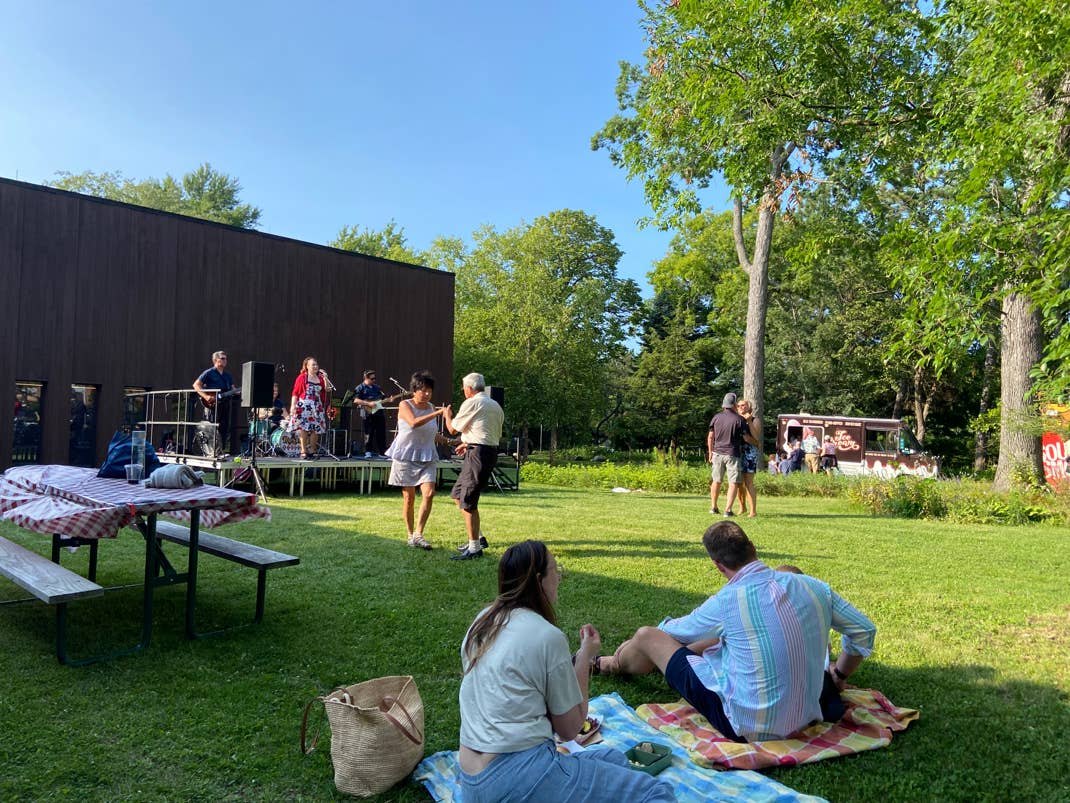 Outdoor park event with a band on stage, people on blankets, and a food truck among trees on a sunny day.
