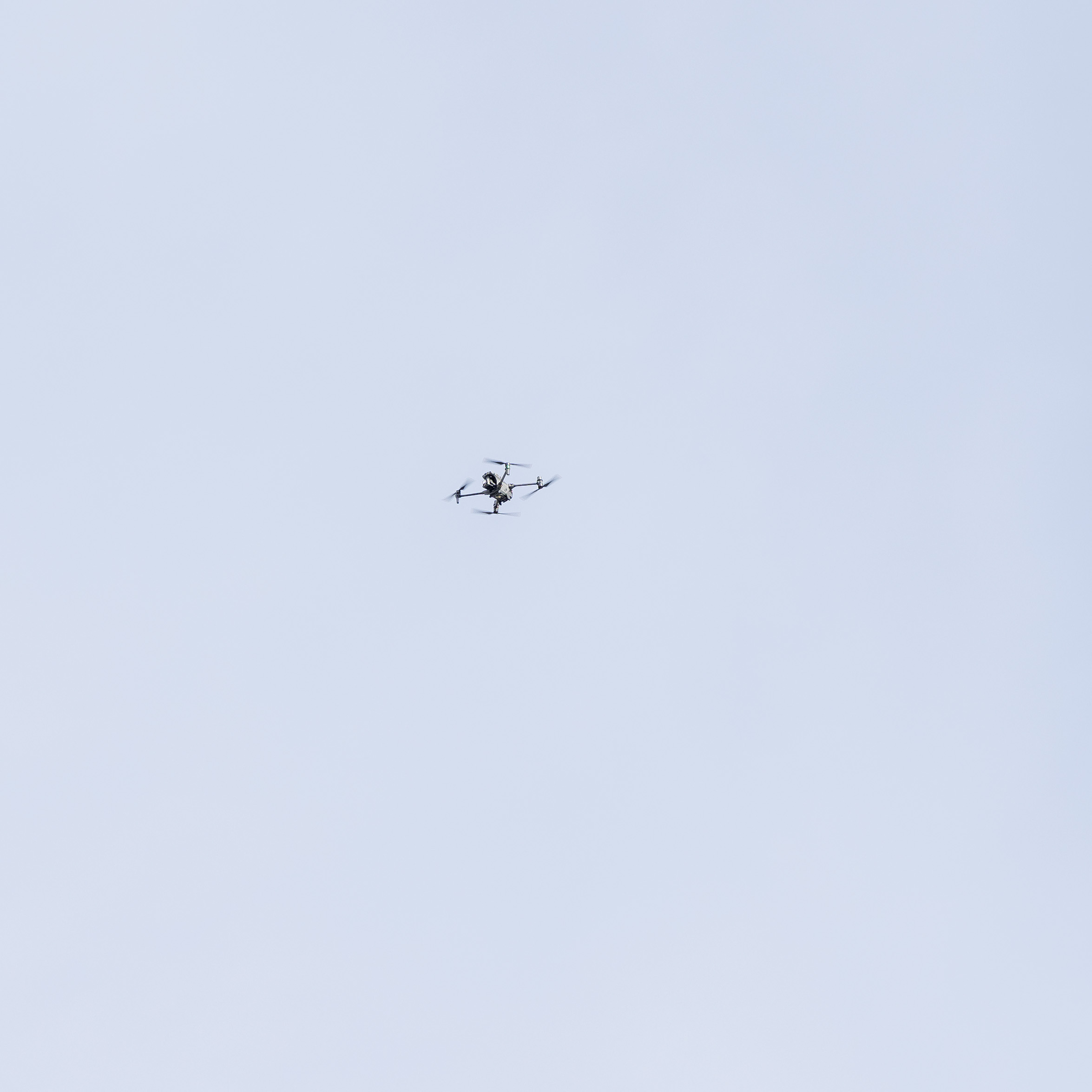 Small multirotor drone flying against an overcast grey sky.