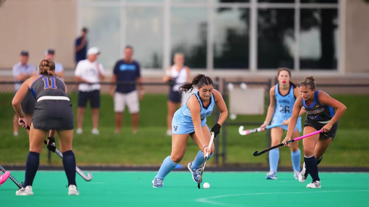 Female field hockey players in light blue uniforms compete on a green turf field.