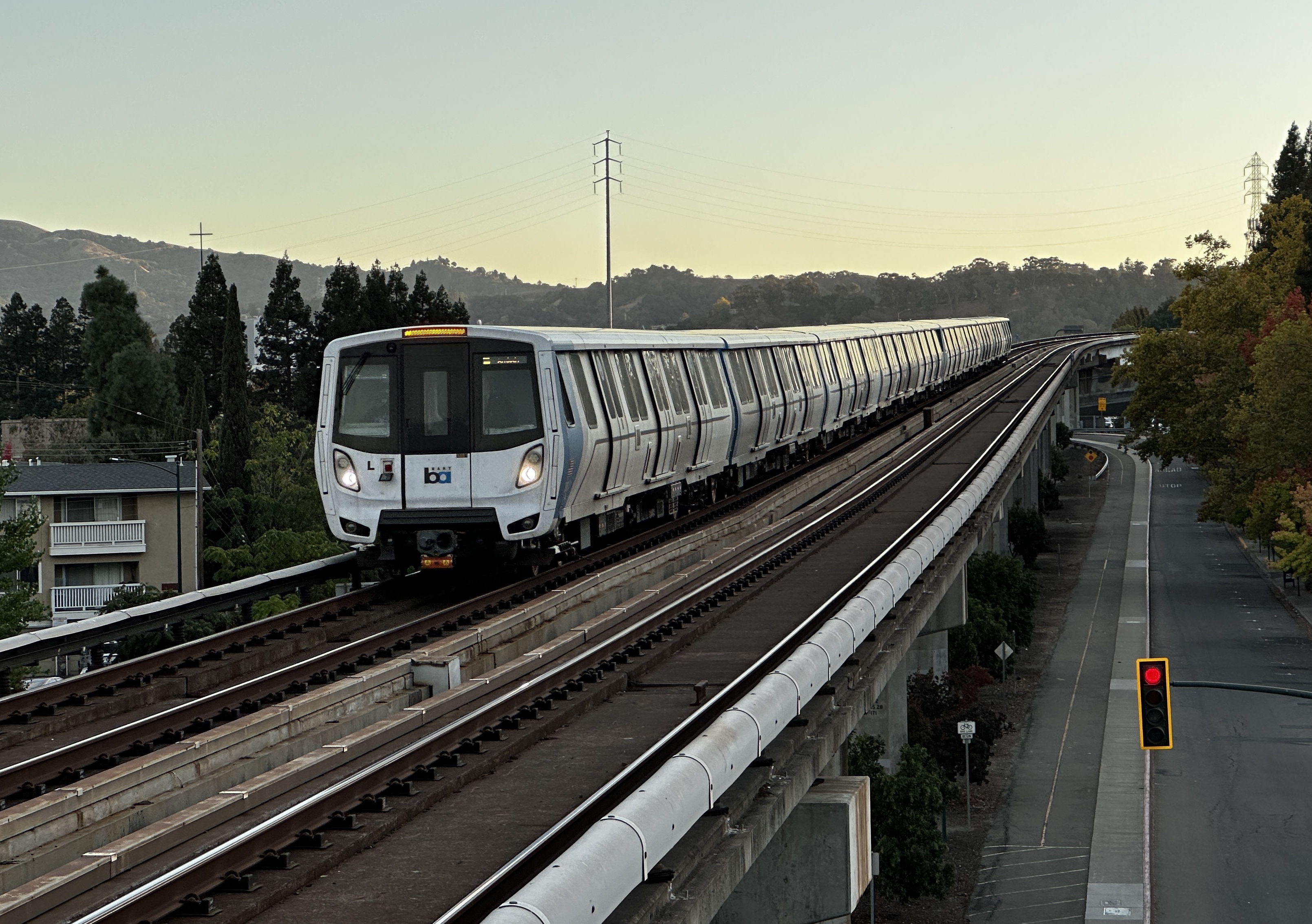 A white BART train travels on an elevated track toward a station, with trees and hills visible.