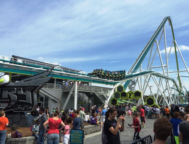 Crowds gather near the entrance of a tall teal steel roller coaster at an amusement park.