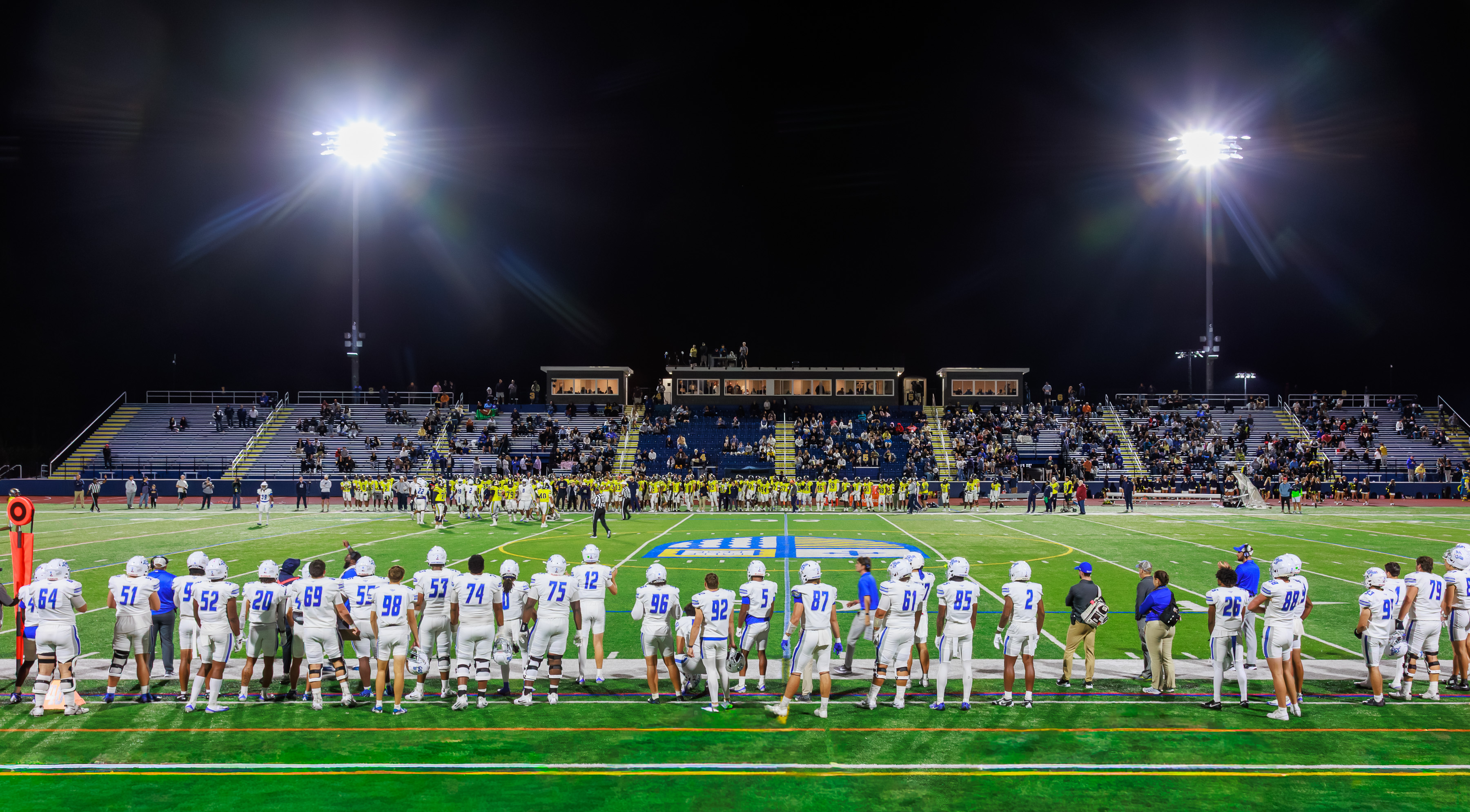 Football players on the sideline of a brightly lit stadium at night with spectators visible in the stands.