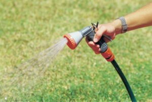 A hand holding a red and gray garden hose spray nozzle watering a green lawn.